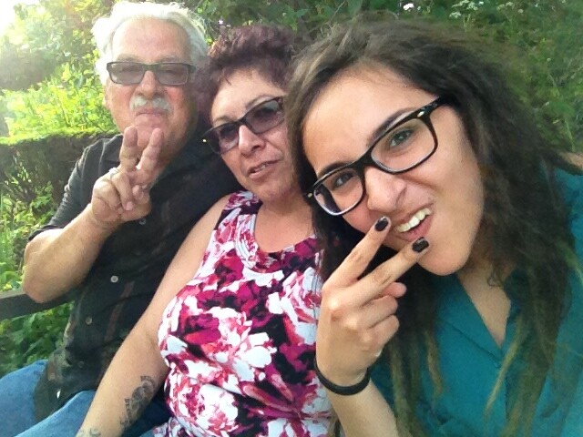 Three people posing outdoors; man with sunglasses left, woman in a floral top center, and a younger woman with glasses on the right flashing peace signs.