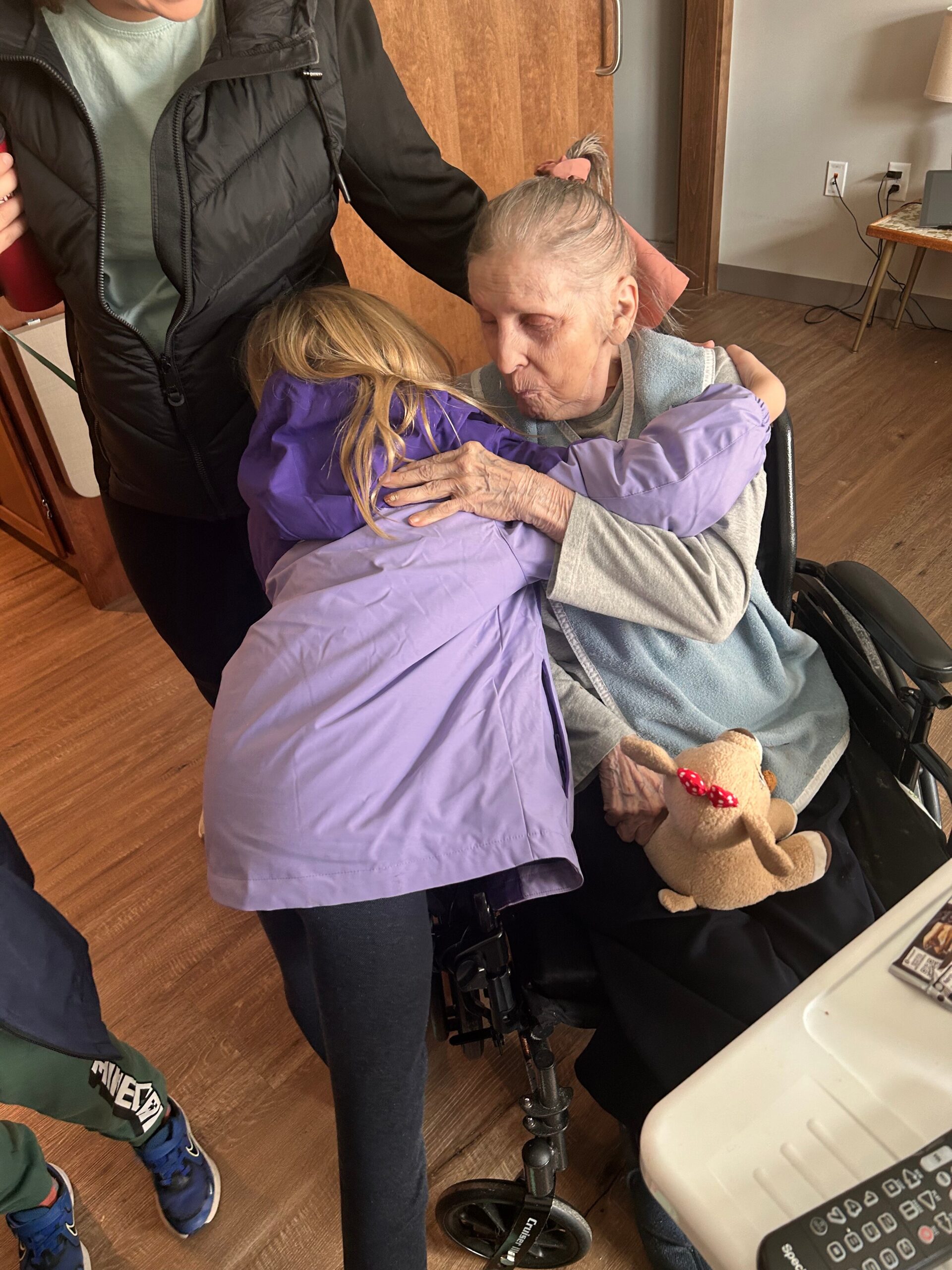Caregiver embracing an elderly woman in a wheelchair, sharing a tender hug in a home setting; teddy bear sits on the woman's lap nearby.