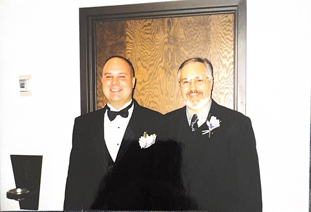 Two men in tuxedos with boutonnieres smiling at the camera, standing in front of a wooden door.