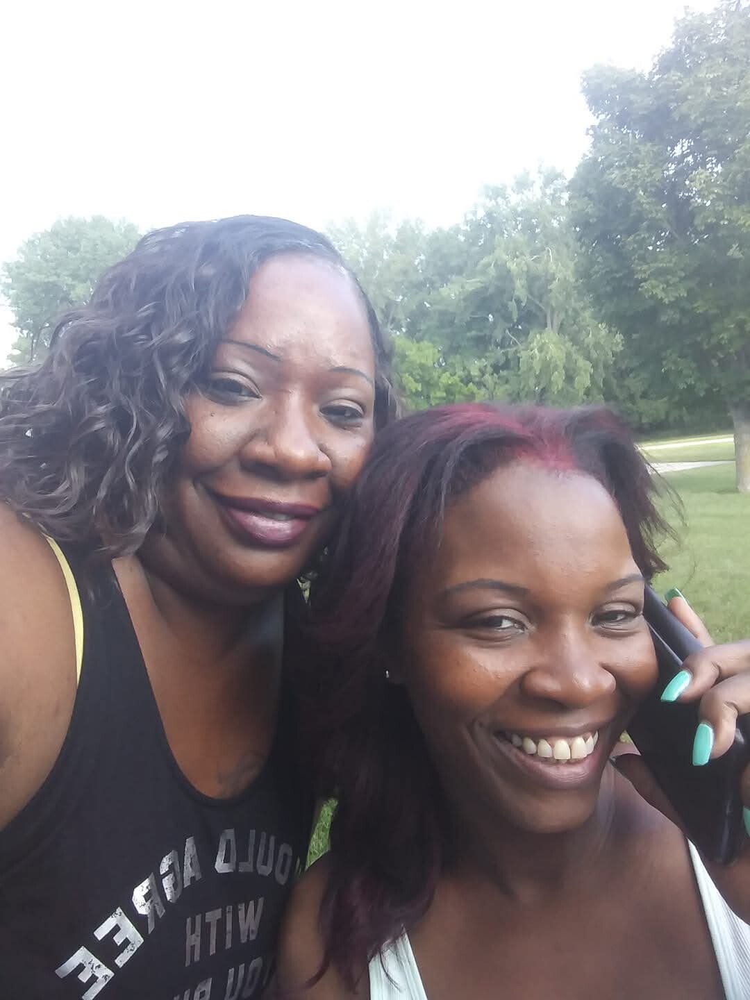 Two smiling women pose for a selfie in a sunny park, one with curly hair in a black tank top and the other with burgundy hair holding a phone to her ear.