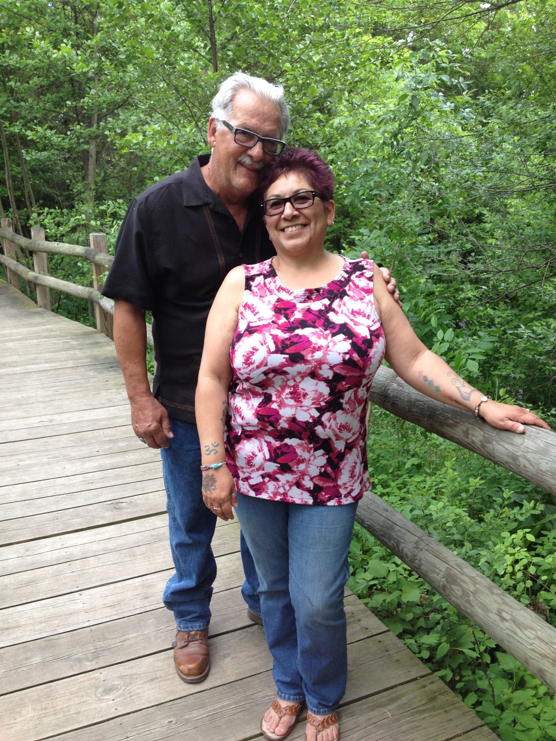 Smiling couple on a wooden boardwalk, surrounded by green foliage; woman wears a pink floral top, man in a black shirt with glasses.