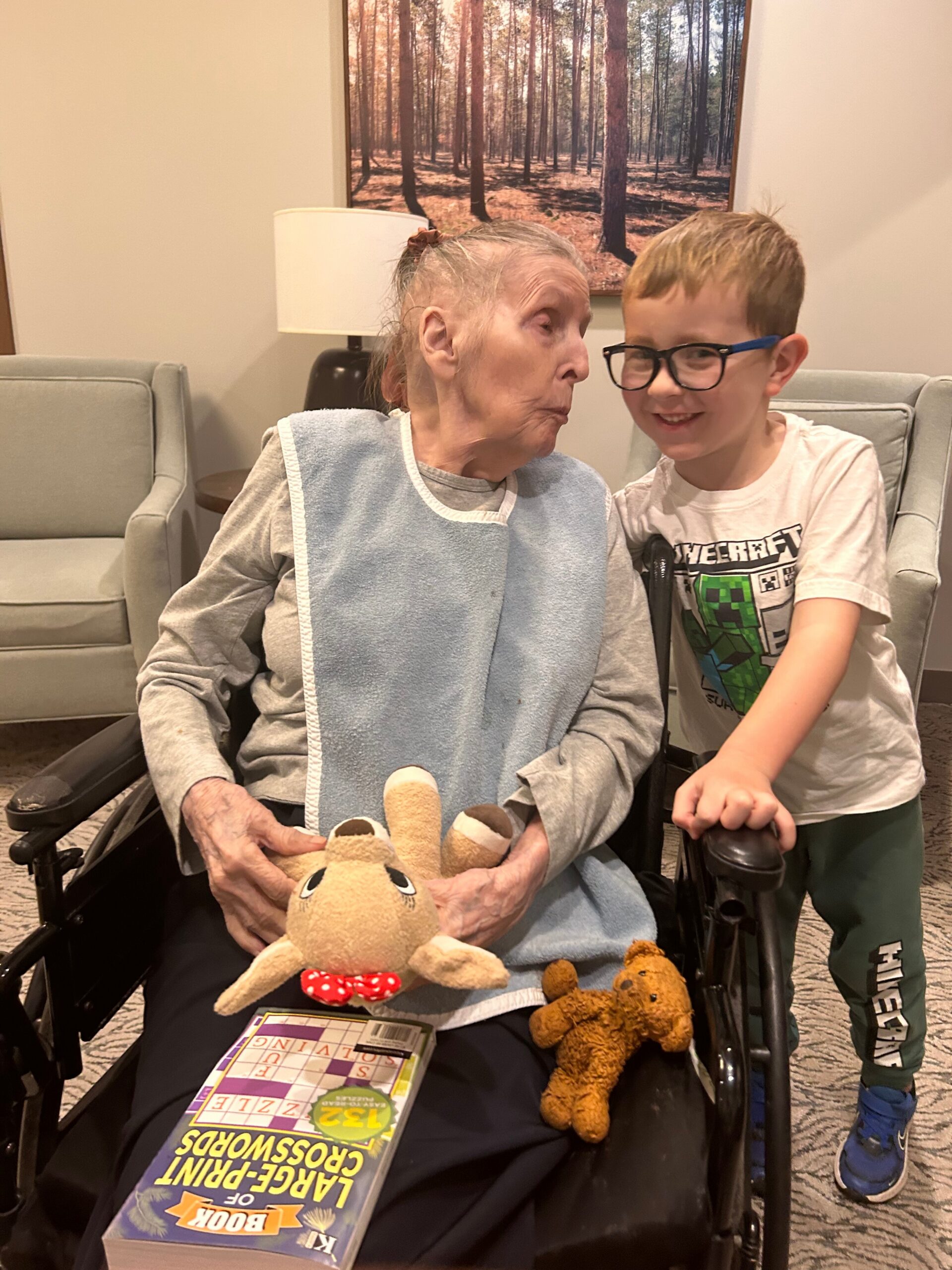 An elderly woman in a wheelchair leans toward a smiling young boy with glasses as they share a moment, with plush toys and a magazine on her lap in a cozy room.