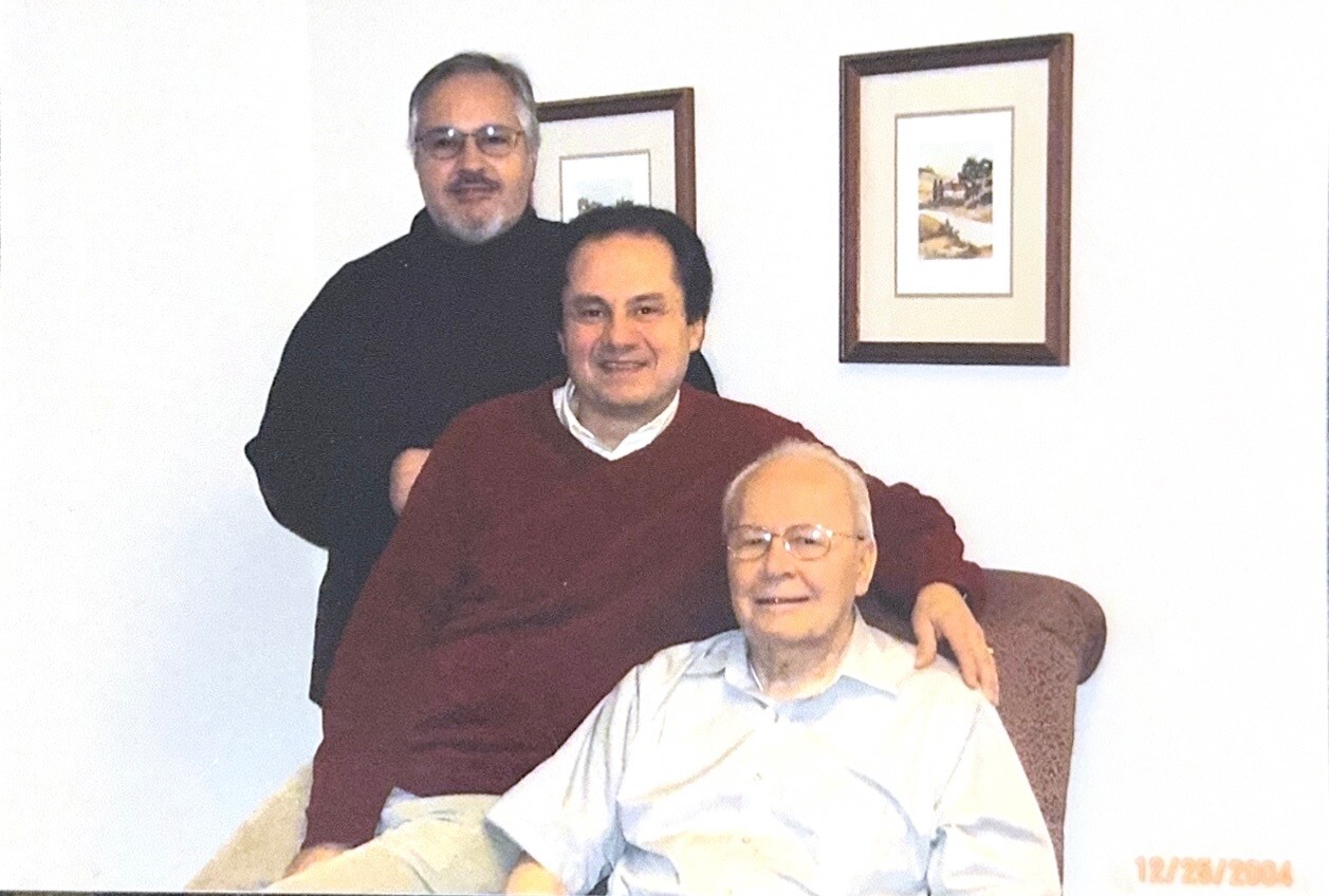 Three men pose in a living room: an elderly man sits in a brown chair, a middle-aged man in a maroon sweater sits beside him, and another man with glasses stands behind them; two framed pictures hang on the wall behind.