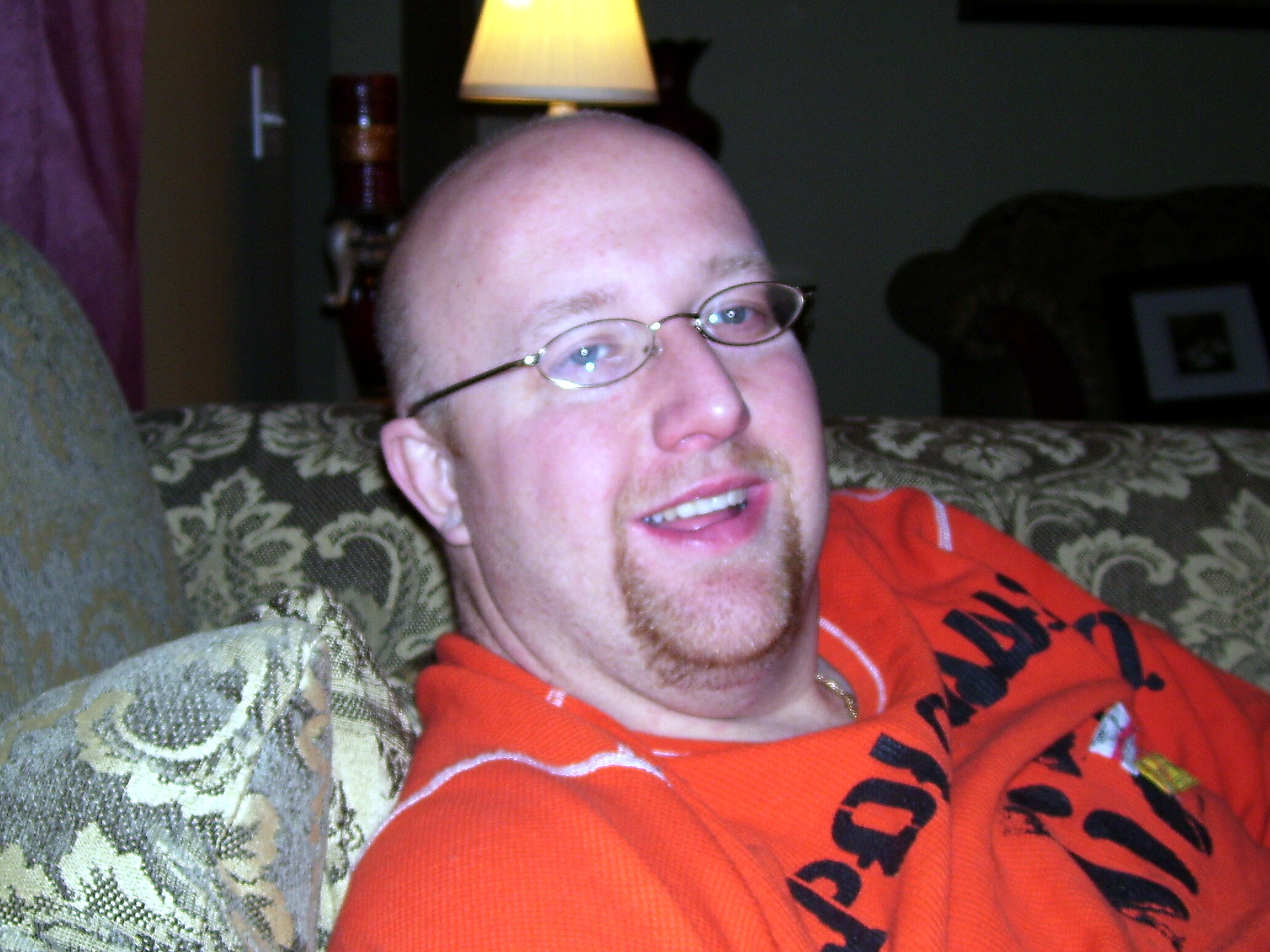 Close-up of a man with glasses lounging on a patterned sofa, wearing an orange shirt and a subtle smile, lamp in background.