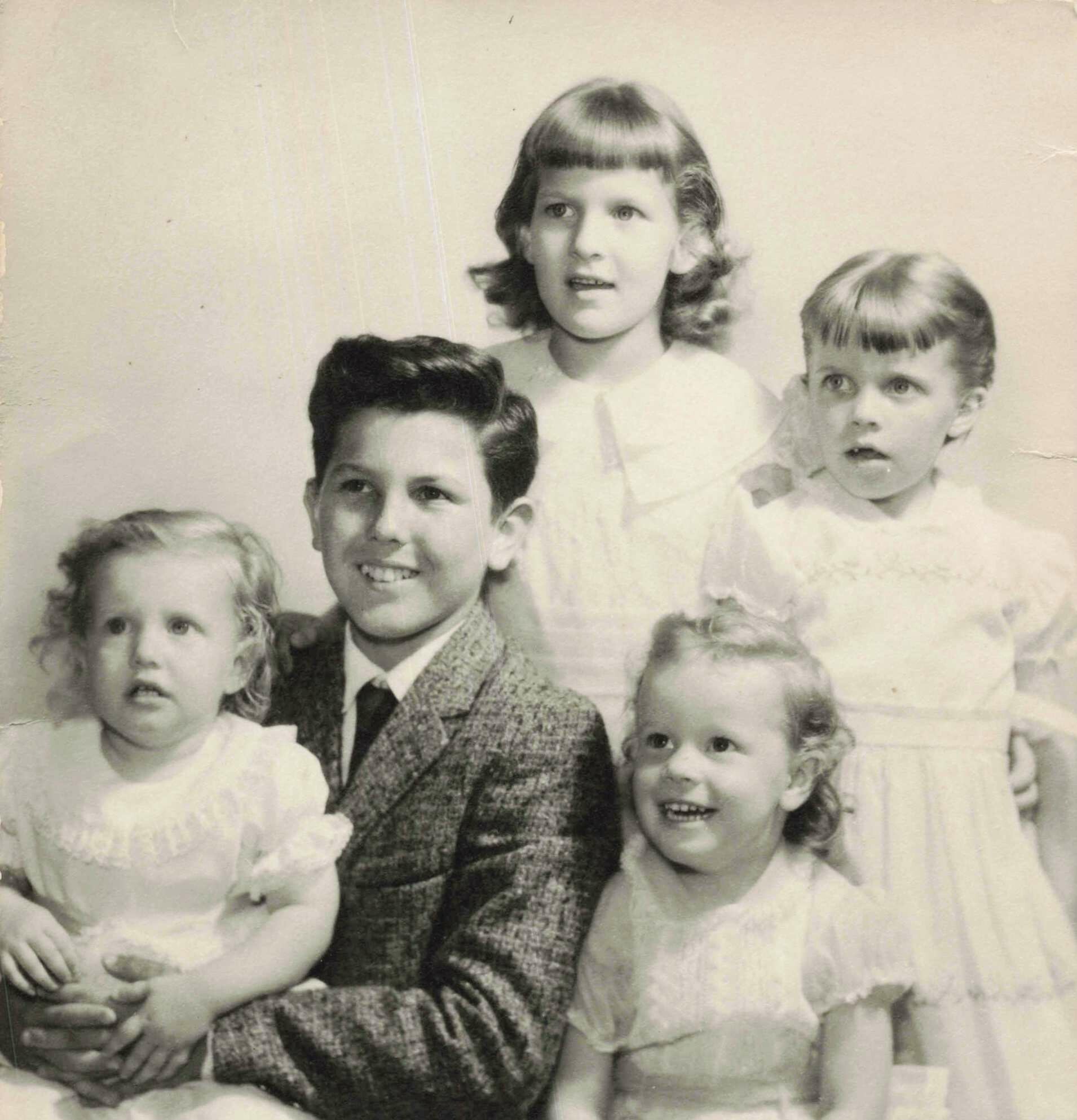 Black-and-white portrait of several children, likely siblings, posing together in vintage clothing with smiles around a central boy in a patterned suit.