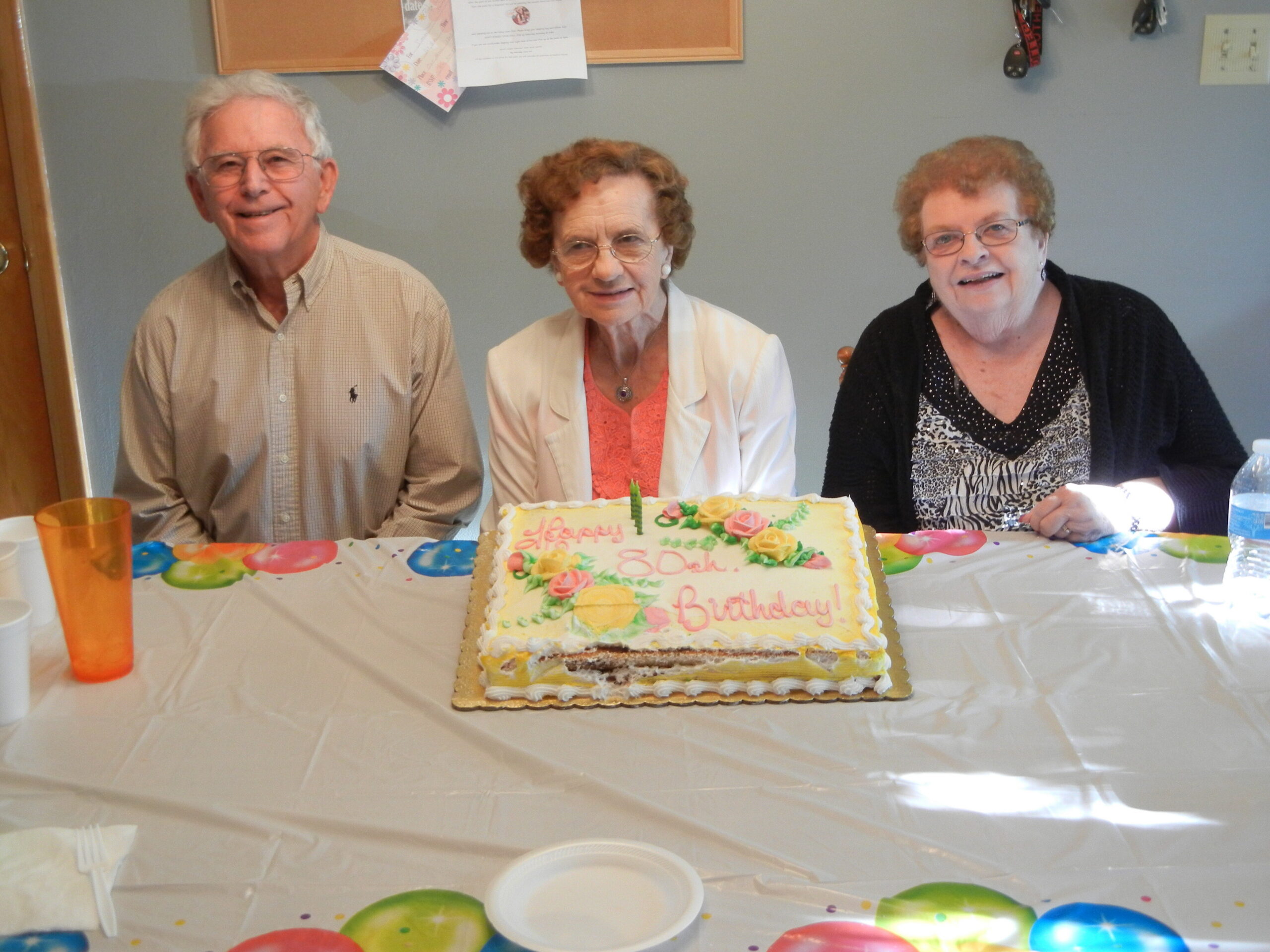 Three elderly adults sit at a table with a decorated birthday cake in front of them, smiling at the camera.