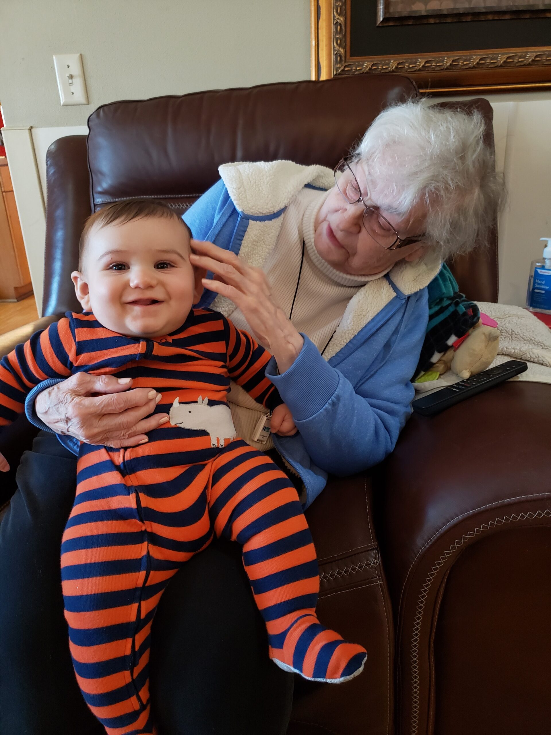 Smiling baby in orange and navy striped outfit sitting on an older woman’s lap on a leather chair; love-filled moment.