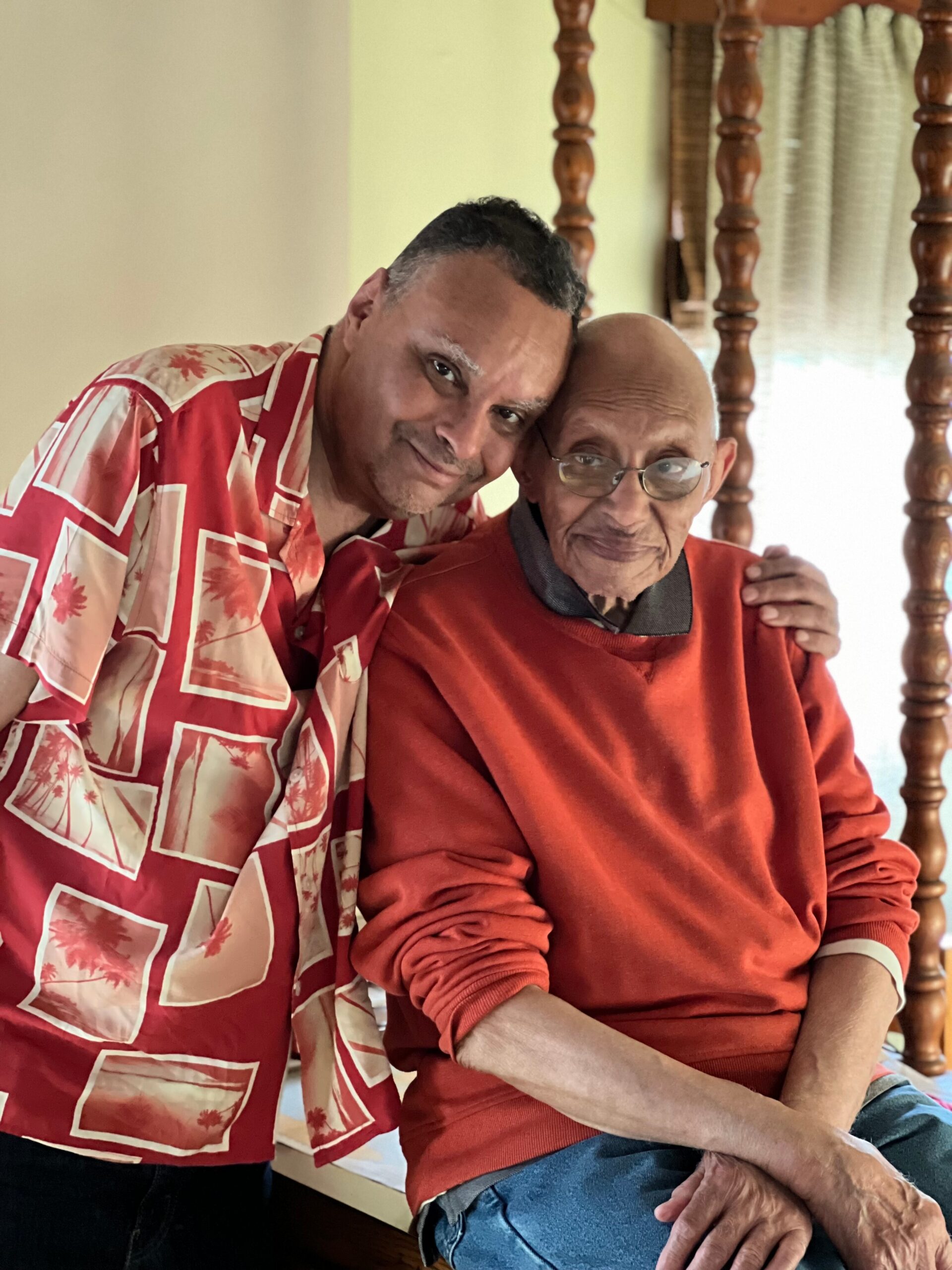 Two men sit closely together indoors; younger man in a red patterned shirt rests his head on an older man in an orange sweater, smiling.