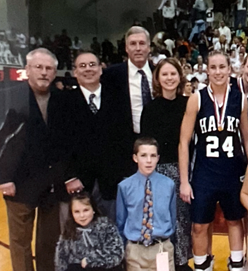 Group portrait on a basketball court: a smiling female player in a navy Hawks jersey #24 with a medal, flanked by three men in suits and two children.