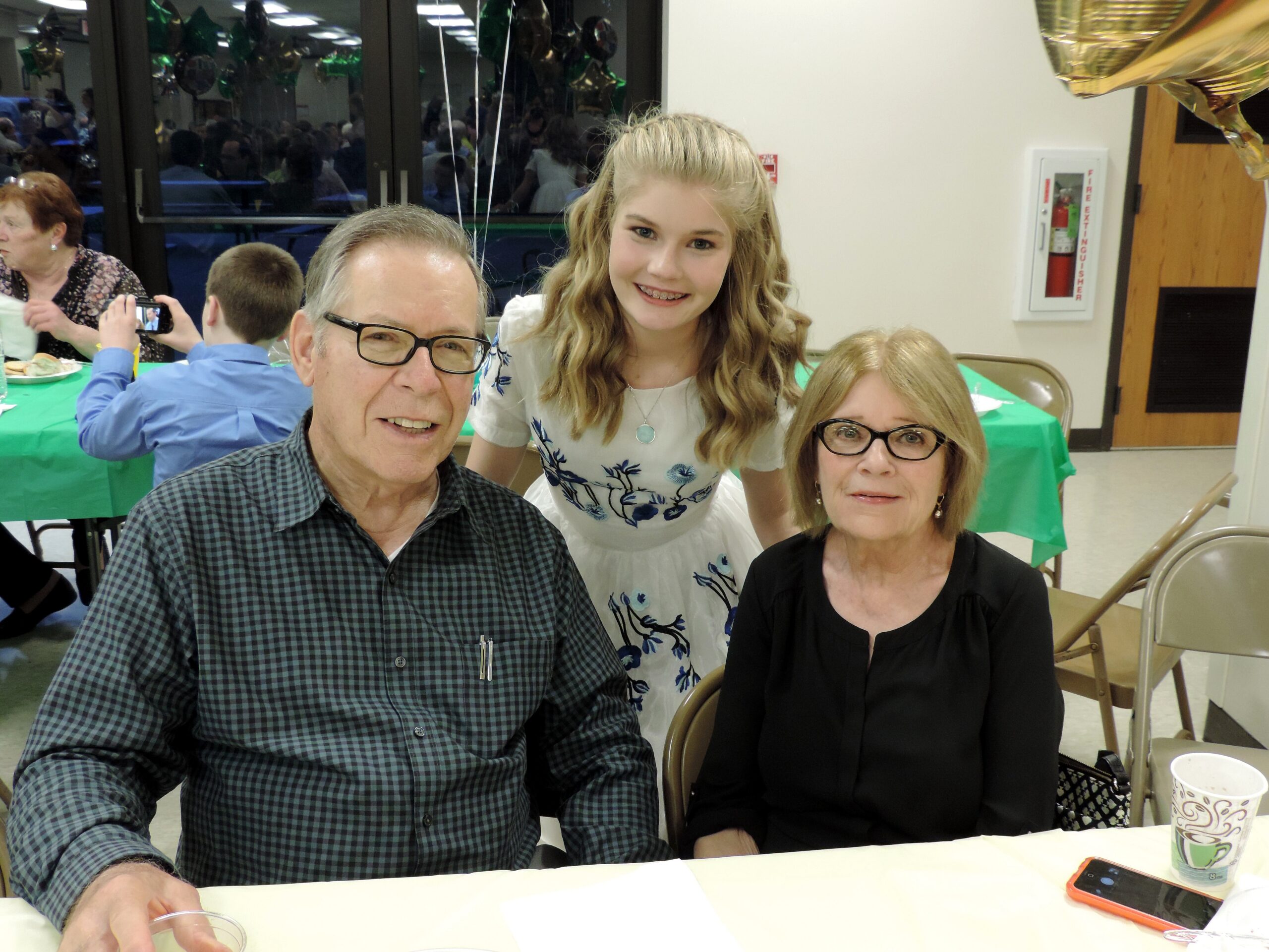 Three people pose for a photo at a party: an older man with glasses, a teen girl in a white floral dress, and an older woman in black smiling at the camera in front of a table with cups.