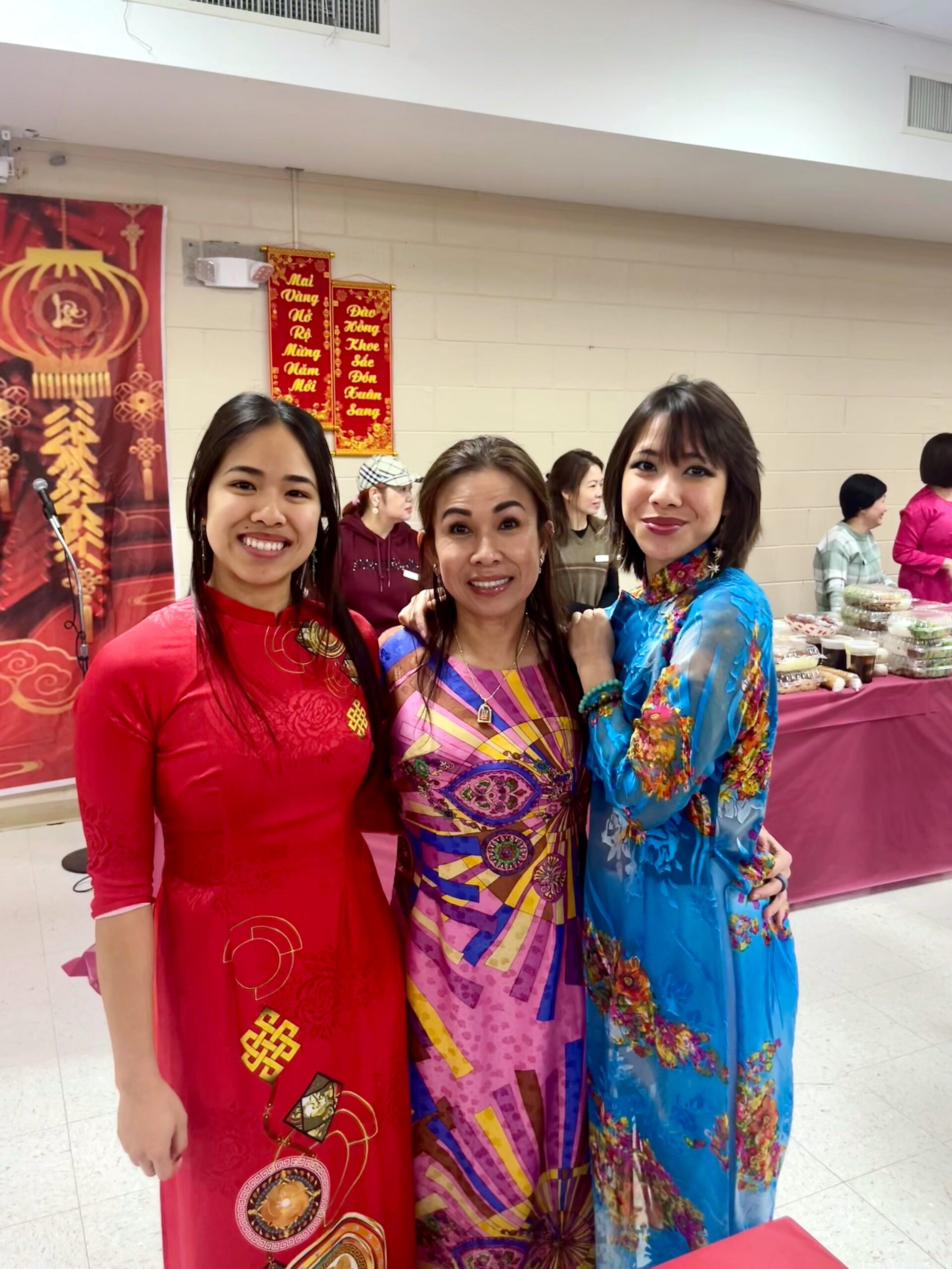 Three women in colorful Vietnamese áo dài posing and smiling at a festive indoor event with red decorations and a table of food in the background.