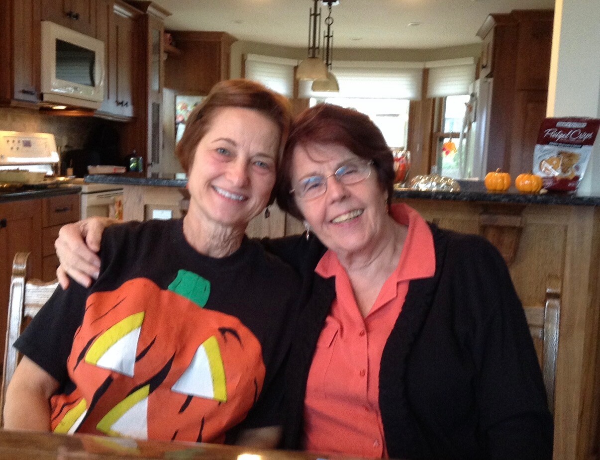 Two smiling women sit close together at a kitchen table, arm around each other, posing for a friendly photo in a home.