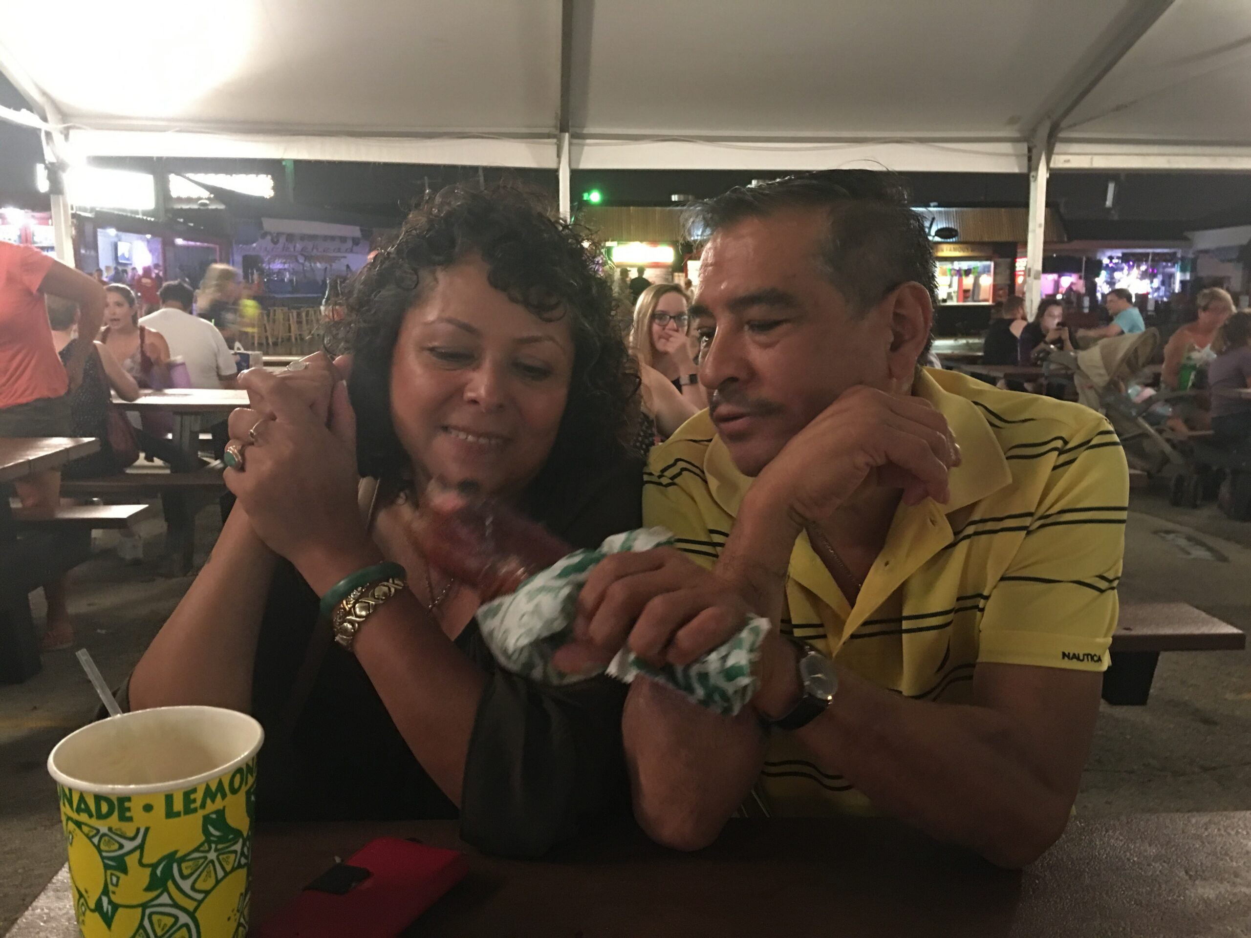 Two people sit closely together at an outdoor night market or festival, sharing a moment and leaning toward each other with drinks in front.