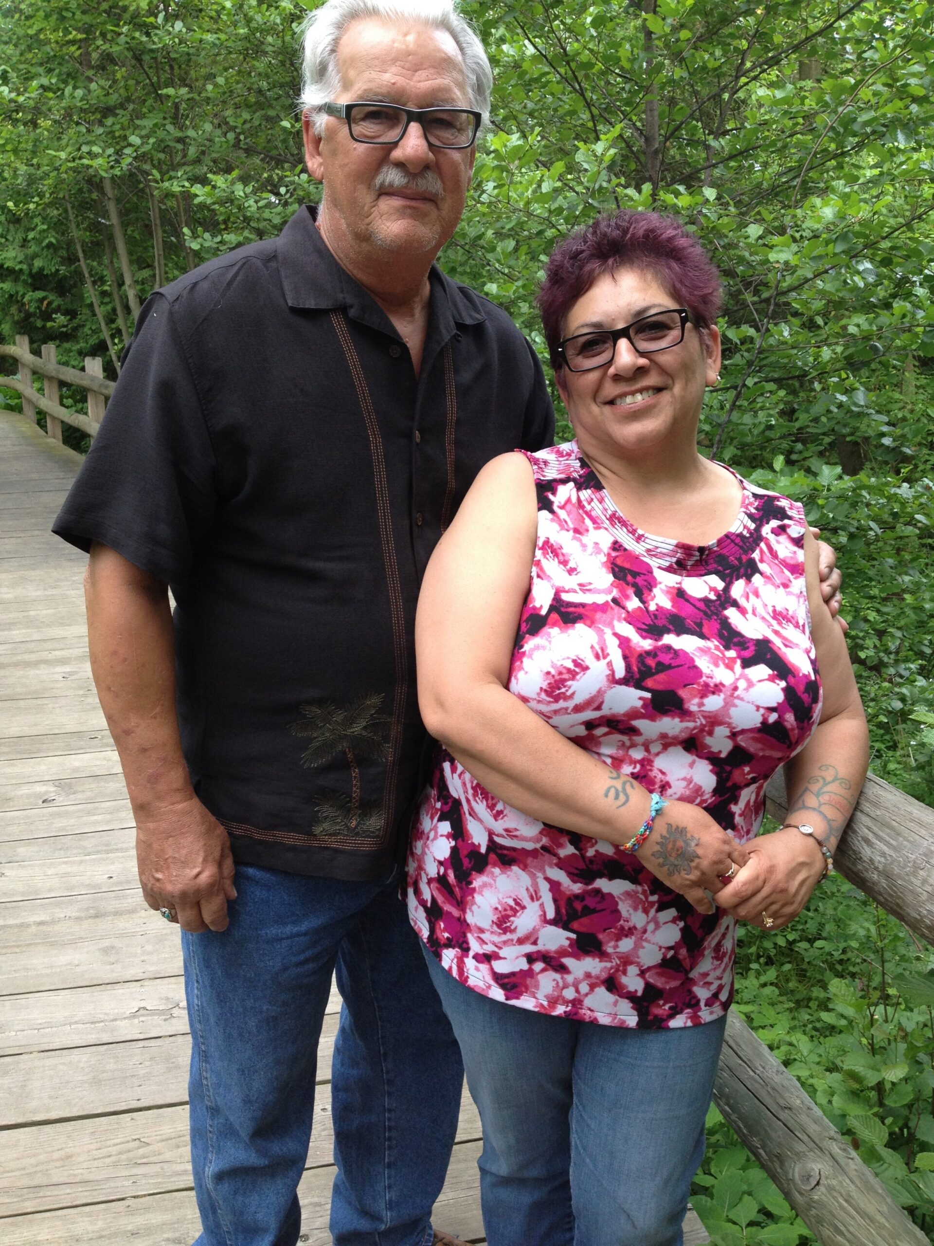 Smiling couple standing together on a wooden path surrounded by green foliage; man in a black shirt and glasses, woman in a pink floral top with short purple hair and glasses.