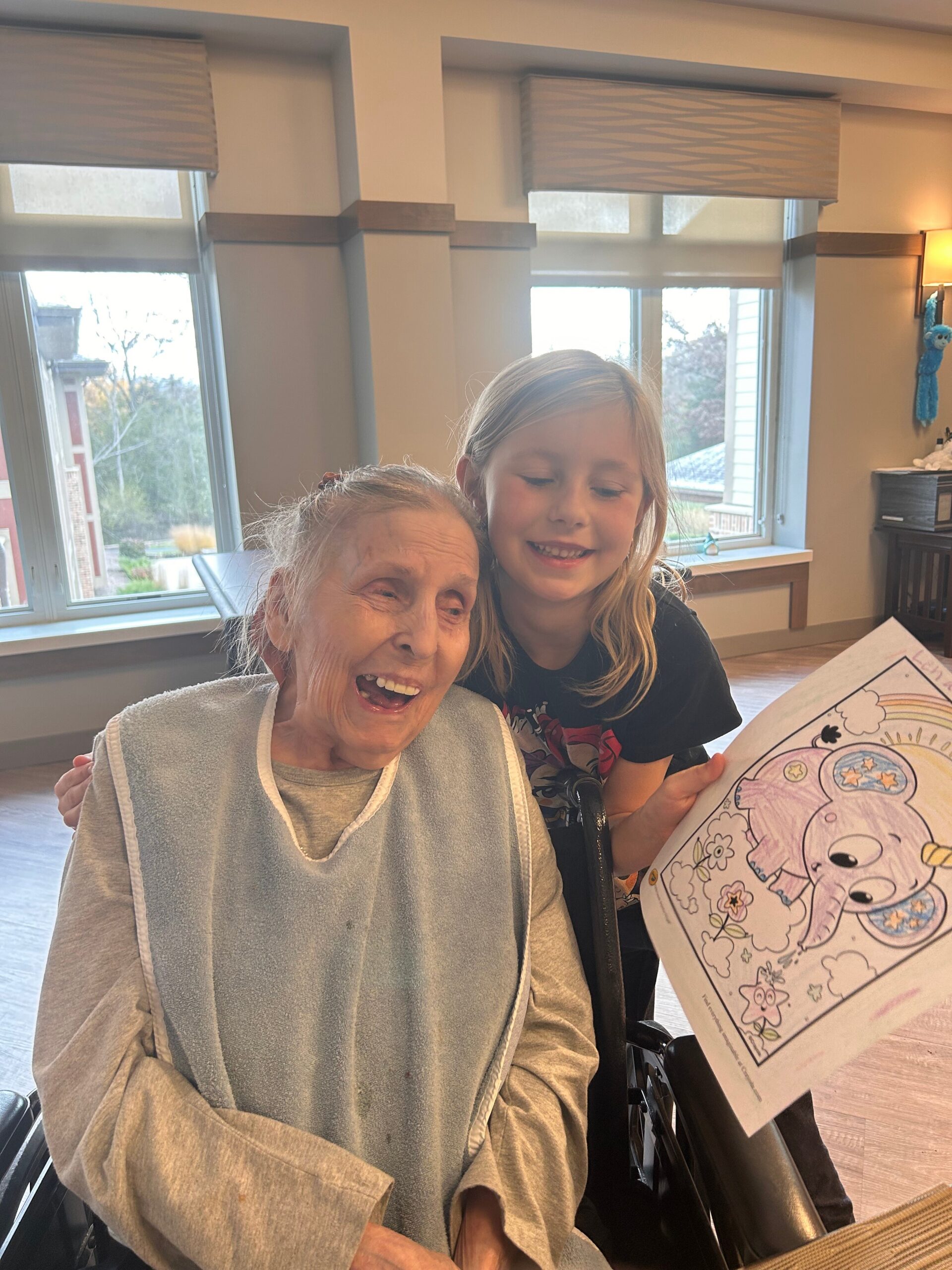 An elderly woman in a wheelchair wearing a bib, smiling as a young girl leans in and shows a coloring book.