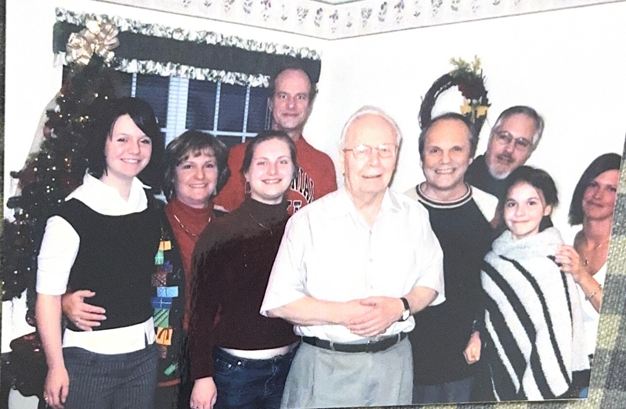 Group photo of eight family members smiling around an elderly man in a white shirt at a holiday gathering in a living room with a Christmas tree in the background.