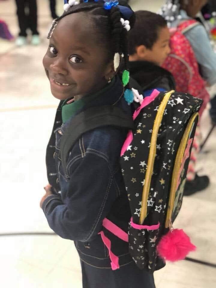 Smiling young girl with braided hair wearing a star-pattern backpack and colorful beads, looking back at the camera in a hallway.