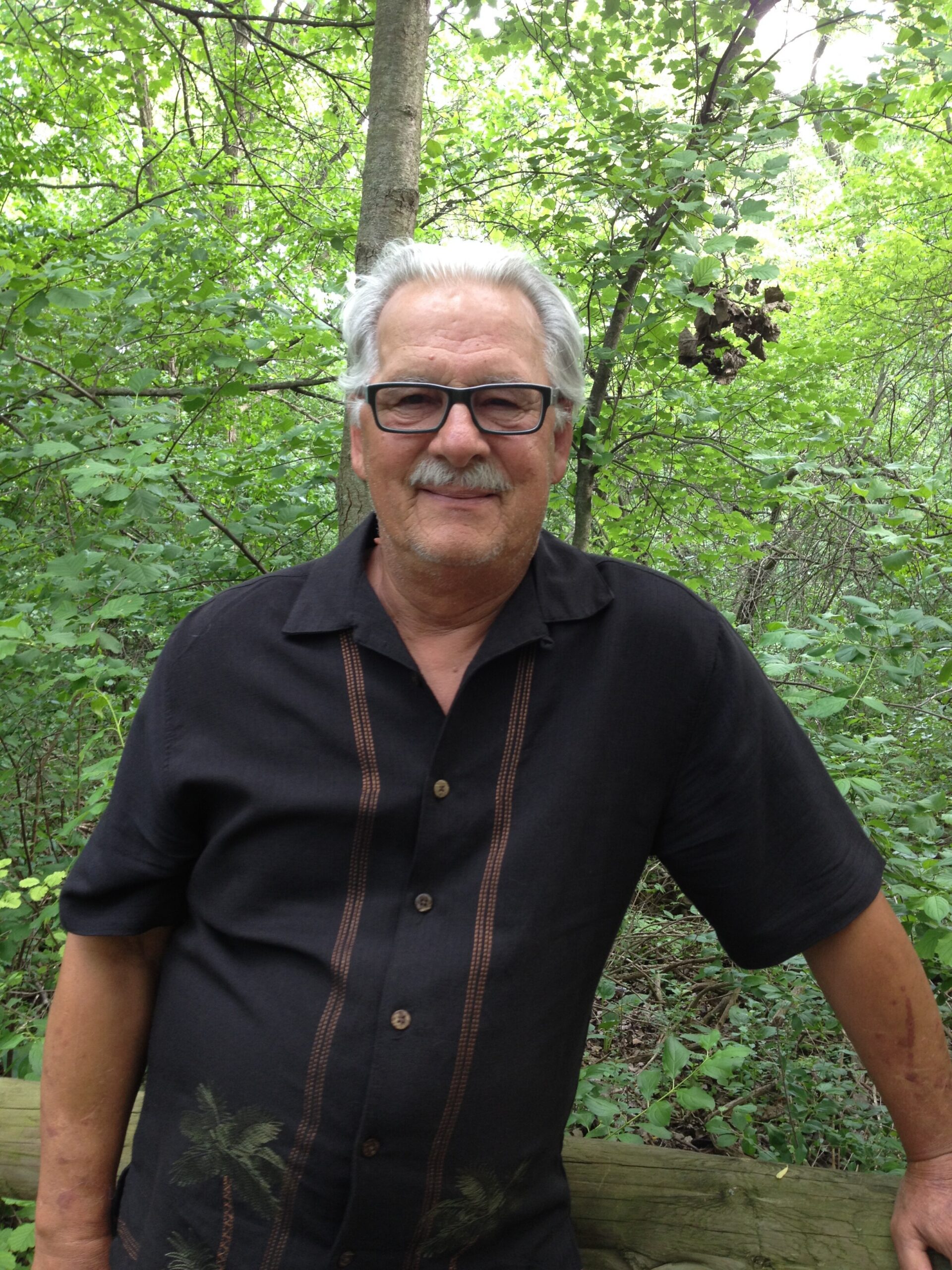 Smiling older man with gray hair and black glasses standing in a green forest. (informative)