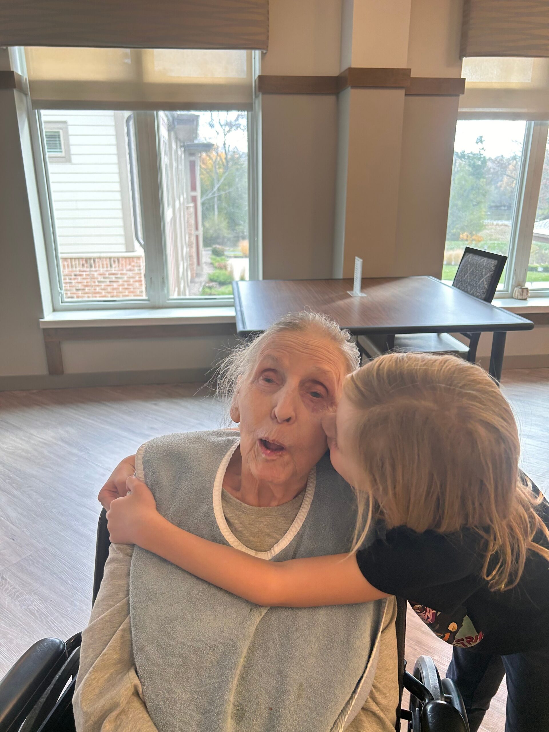 Caregiver shares a warm hug with an elderly woman in a wheelchair, inside a bright room by large windows.