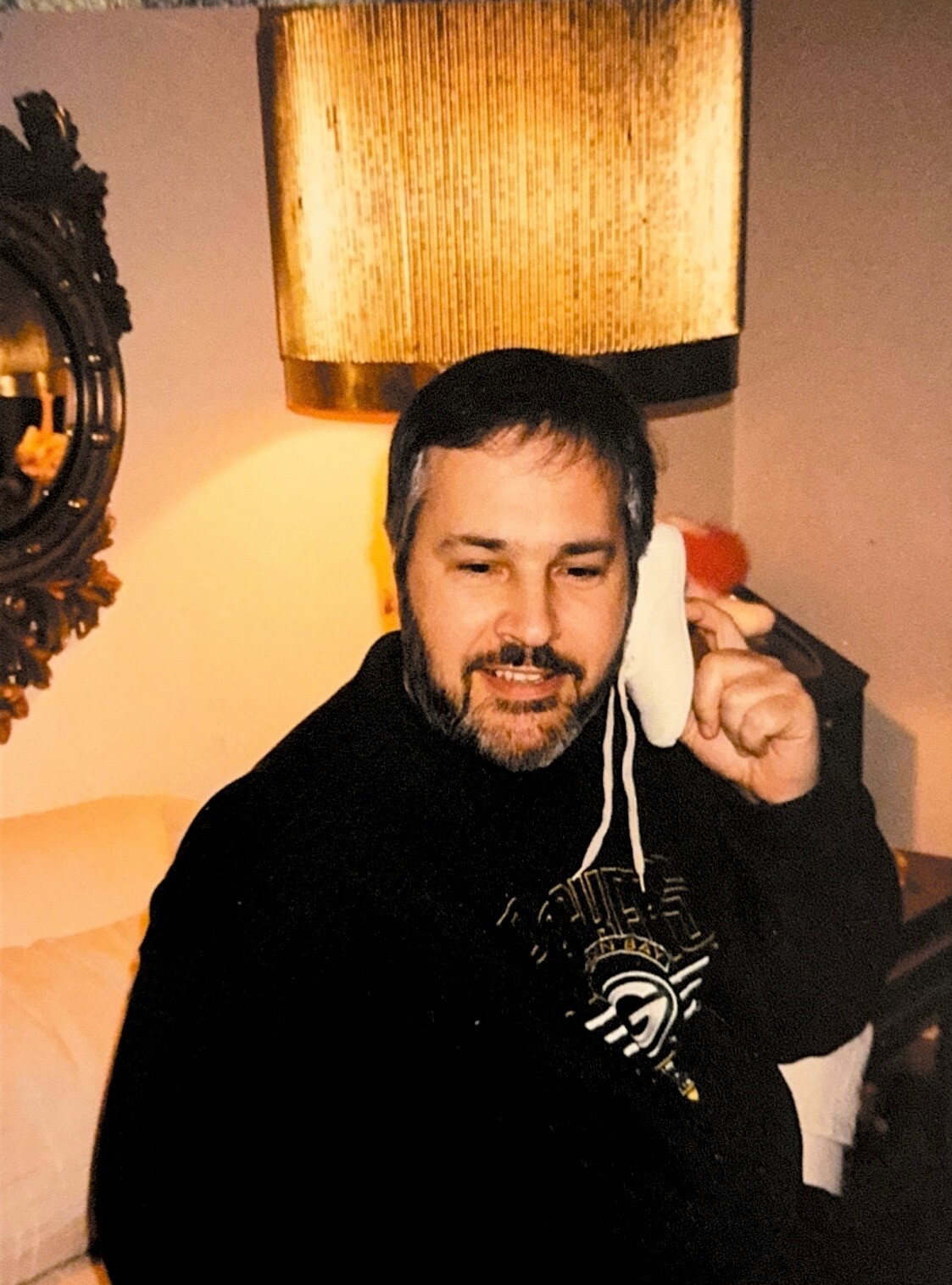 Smiling man with a beard in a black hoodie holds a white sock to his ear, as if talking on the phone, indoors beside a warm lamp and mirror.