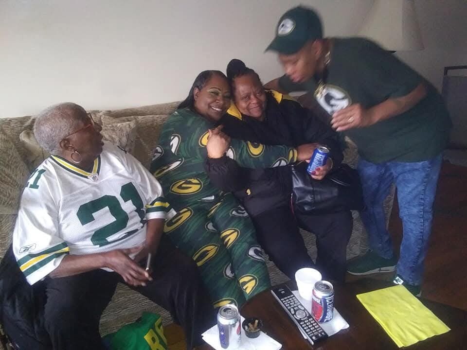 Group of four adults on a couch wearing Green Bay Packers gear, smiling and posing together at a casual gathering.