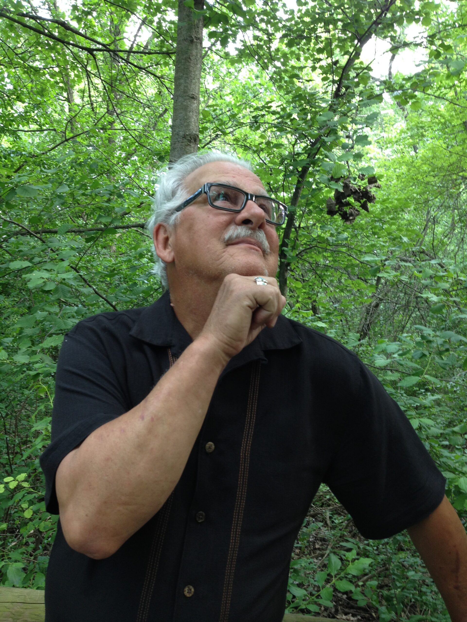 Man with glasses in a black shirt, standing in dense green forest, chin resting on his knuckles, looking upward thoughtfully.