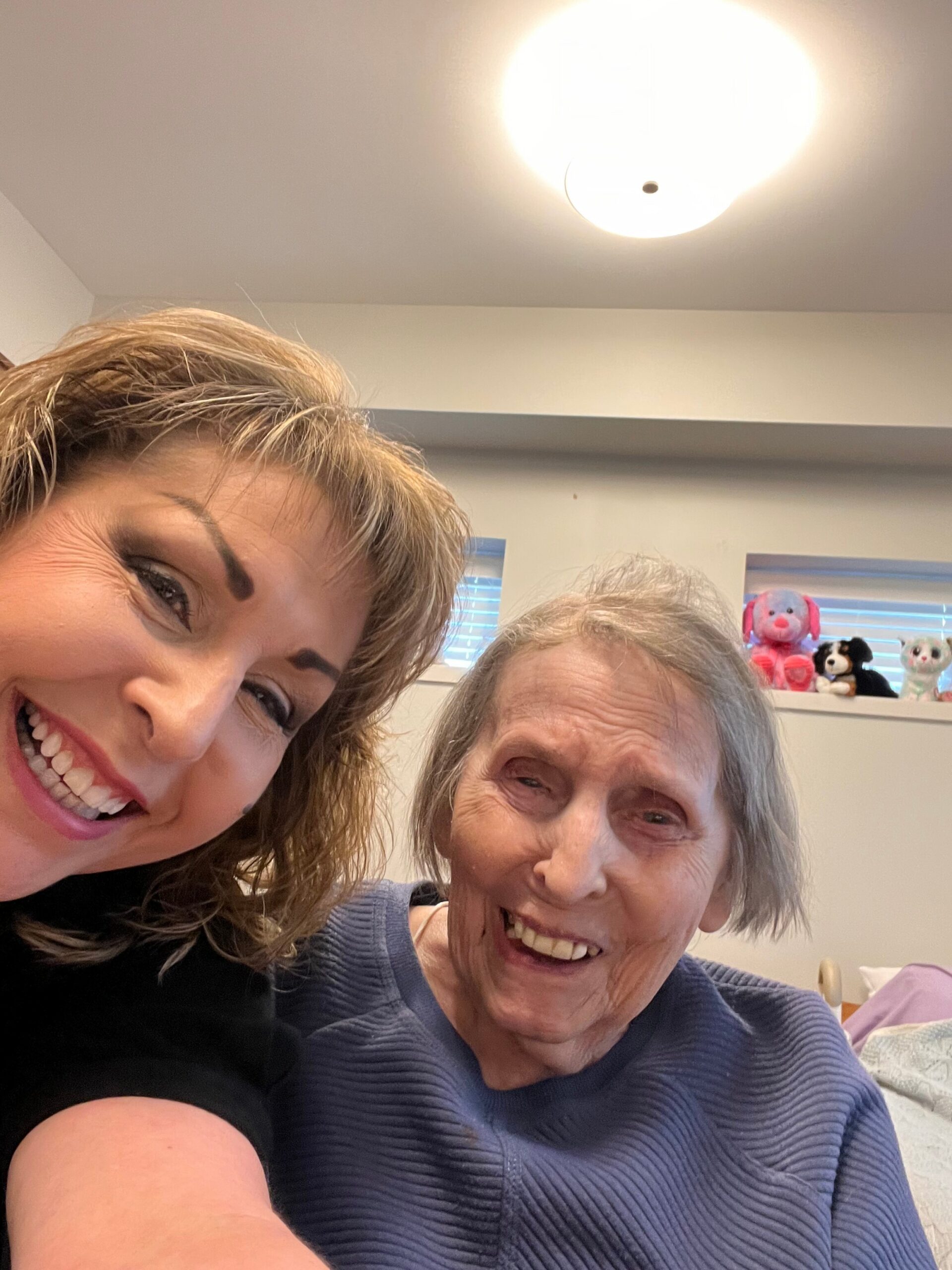 Two women smiling for a selfie indoors, with stuffed animals on a shelf in the background