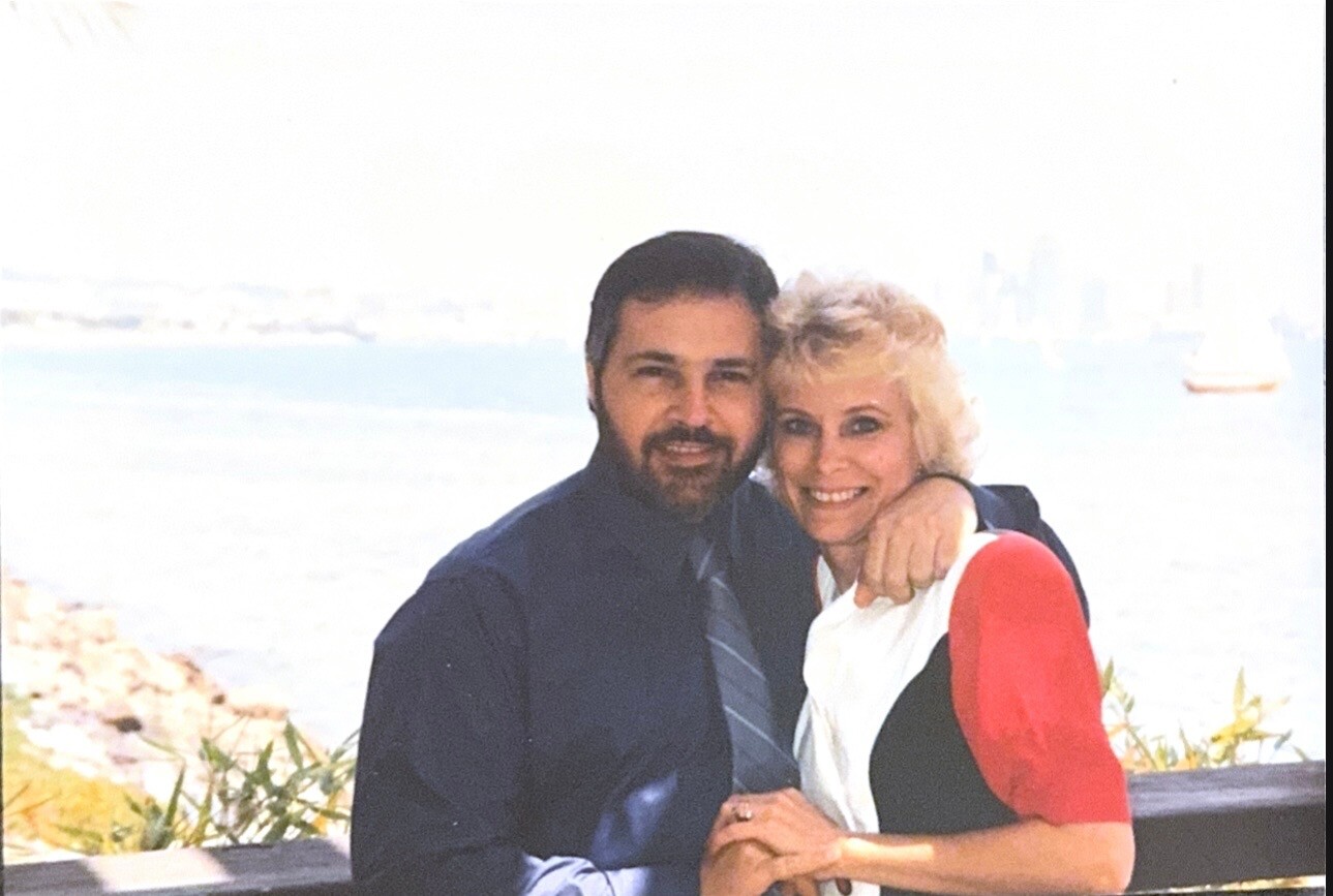 A smiling couple stands arms around each other by the waterfront, posing for a photo.