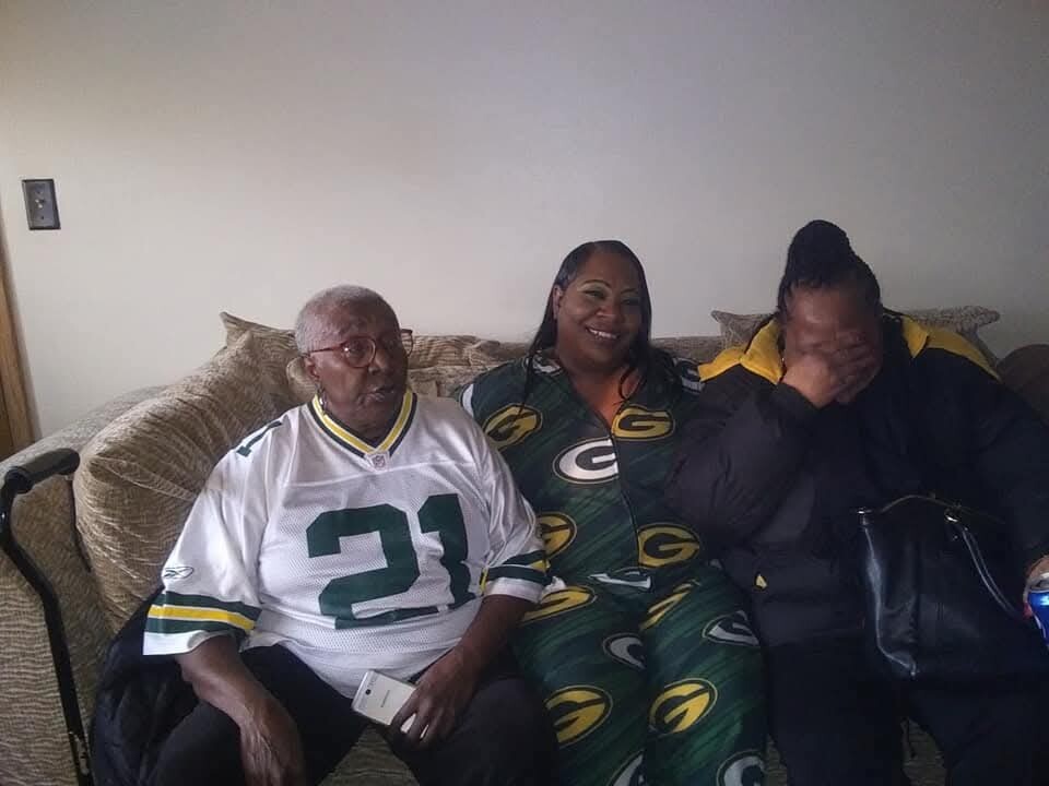 Three people sit together on a beige sofa: an older man in a white Green Bay Packers jersey, a smiling woman in a green Packers dress, and a person in a dark jacket covering their face with their hand.