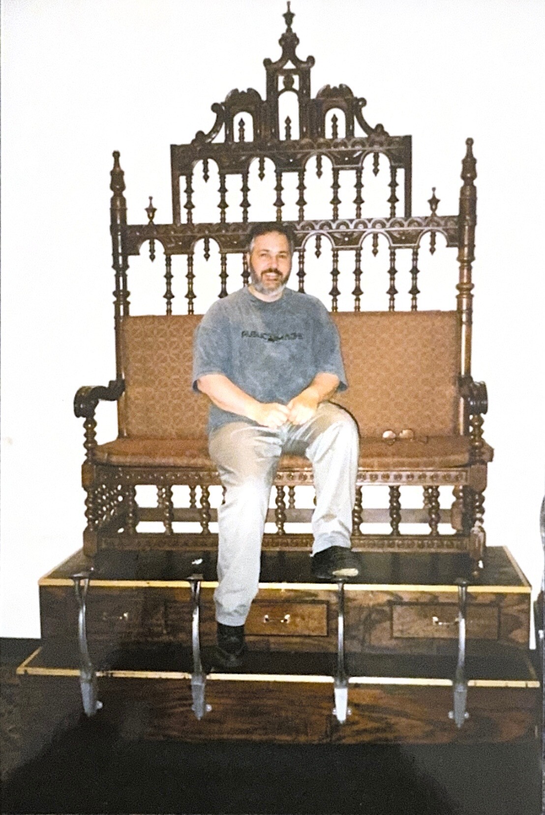 Man sits on an antique carved wooden throne with a tall, decorative back inside a room.