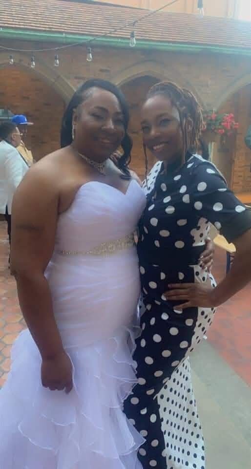 Two women pose together at an outdoor wedding: one in a strapless white wedding gown, the other in a black-and-white polka dot dress, both smiling.