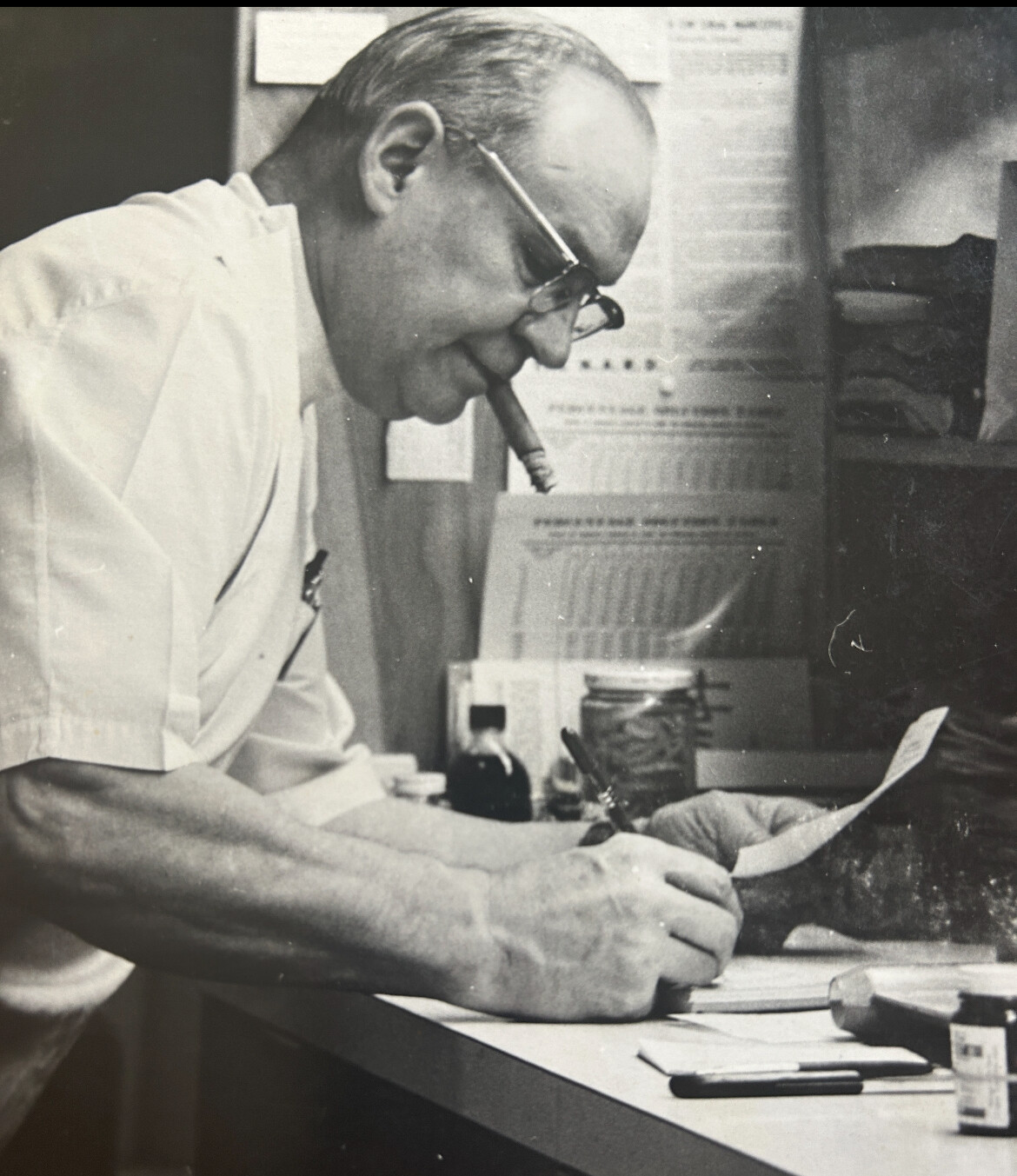 Man with glasses writes at a cluttered desk, cigarette in mouth, focused on paperwork.