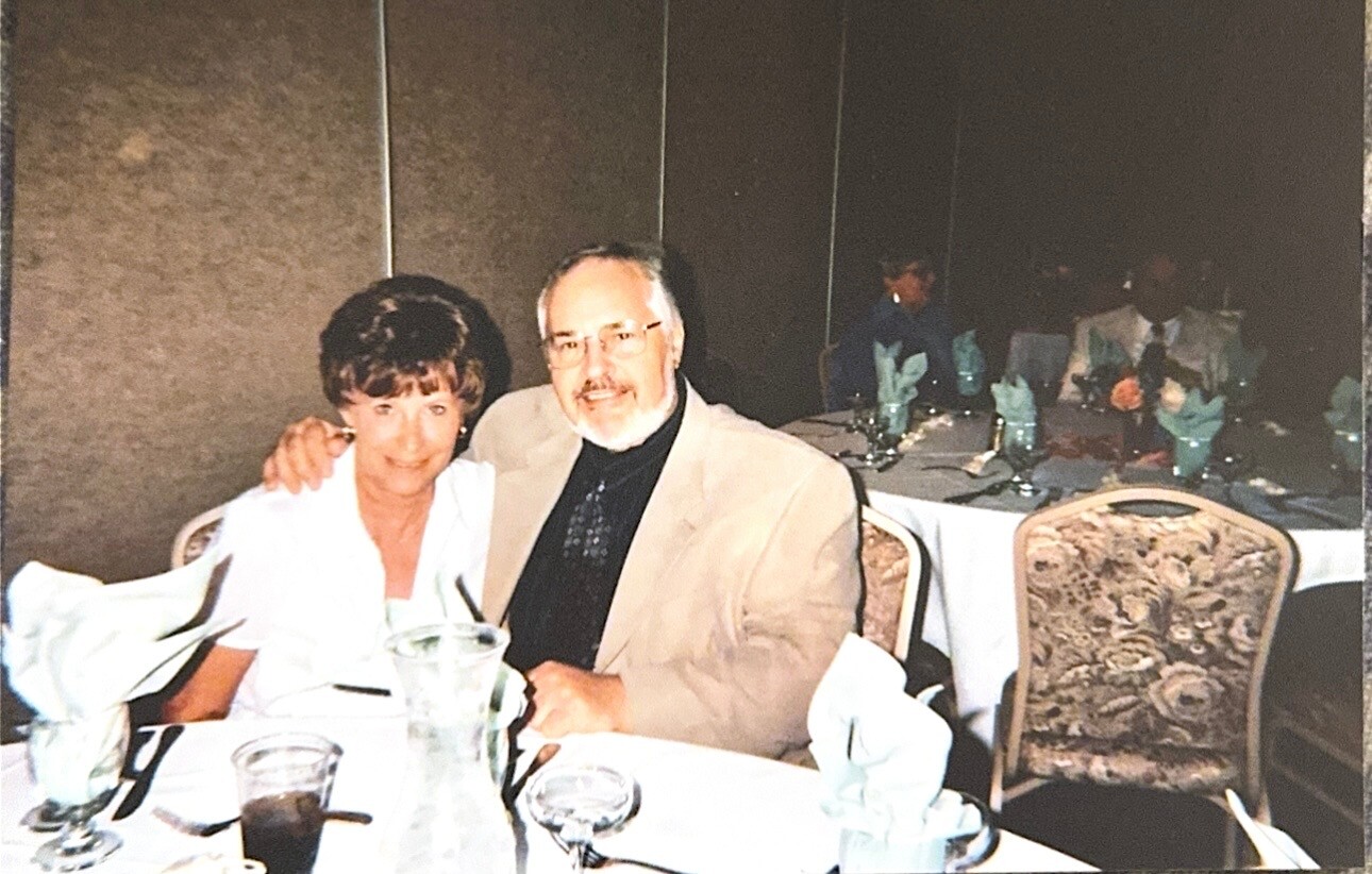 A smiling couple seated at a banquet table with folded napkins and glassware, posing for the camera.