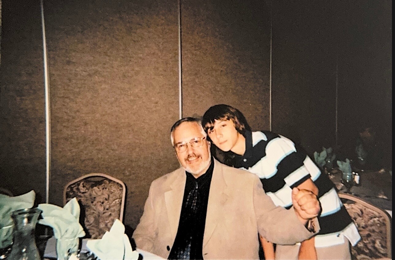 Older man with glasses in a beige blazer sits at a table, younger man in a striped polo leans on his shoulder.