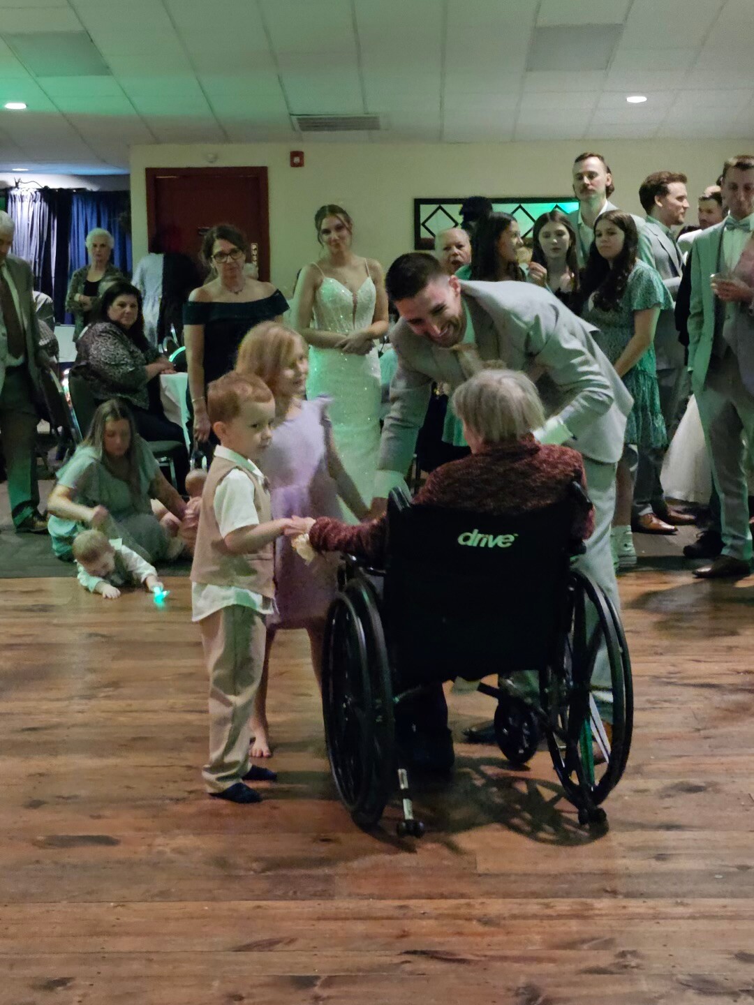 A man in a light suit leans toward a woman in a wheelchair as children stand nearby at a formal event.