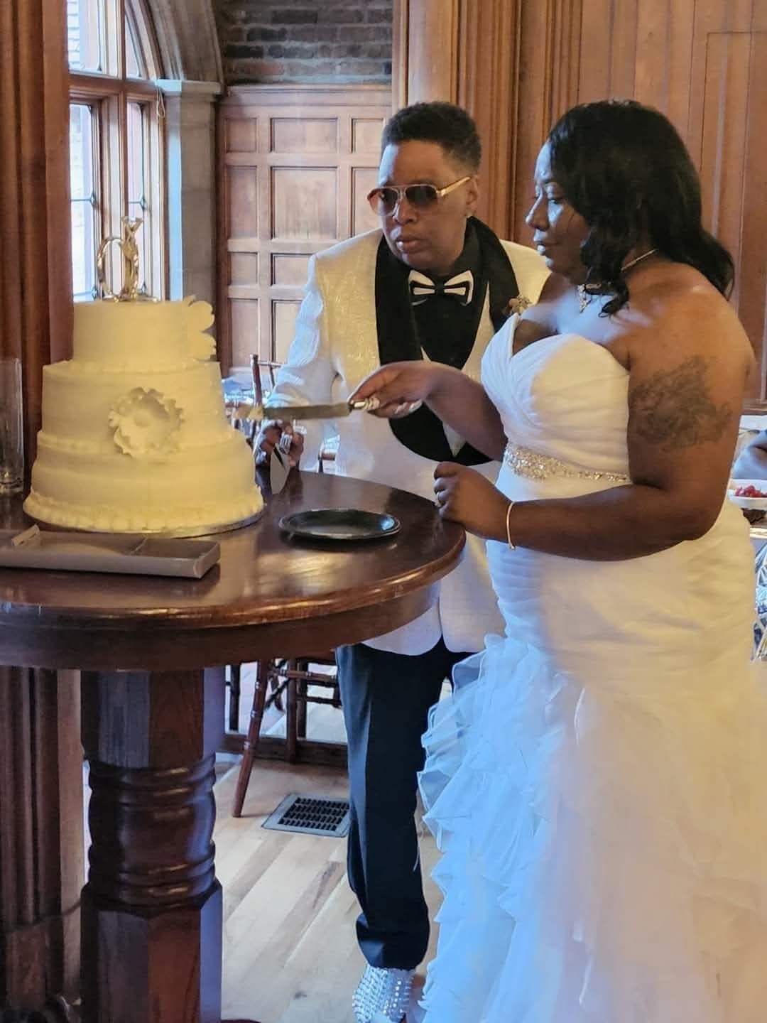 Bride in a white gown and groom in a white suit cut a three-tier wedding cake together at a wooden table.