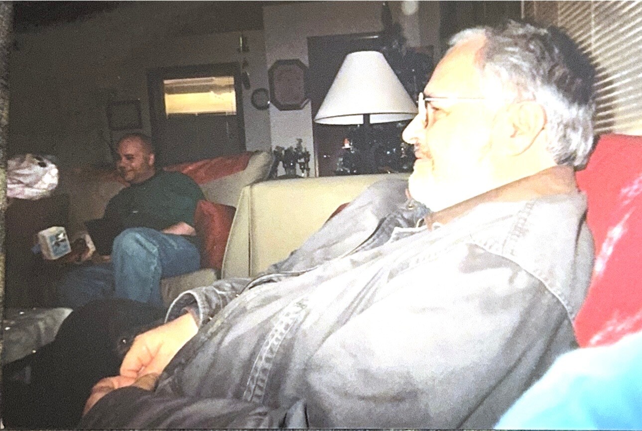 Two men relax in a cluttered living room; foreground man in denim shirt and glasses, background man on the sofa with a mug.