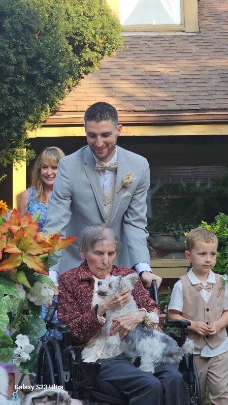 A man in a light gray suit helps an elderly woman in a wheelchair pet a small white dog at a garden wedding, with a boy nearby and flowers nearby.