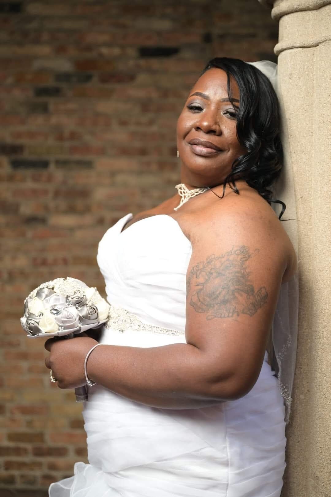 Bride in a strapless white wedding gown, holding a bouquet of white roses, leaning against a stone column beside a brick wall.