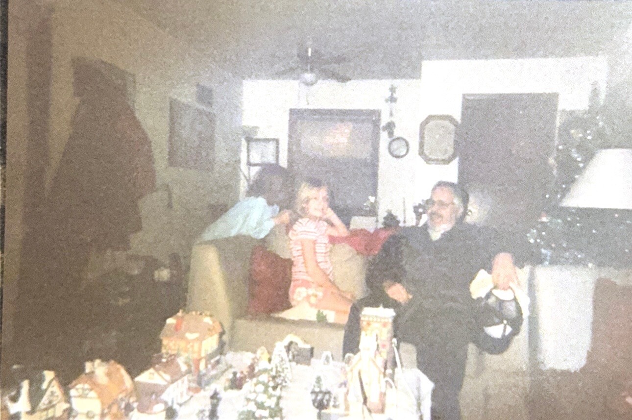 Family gathered in a living room around a table of miniature Christmas village displays; a child in a striped dress sits on a couch beside adults watching.
