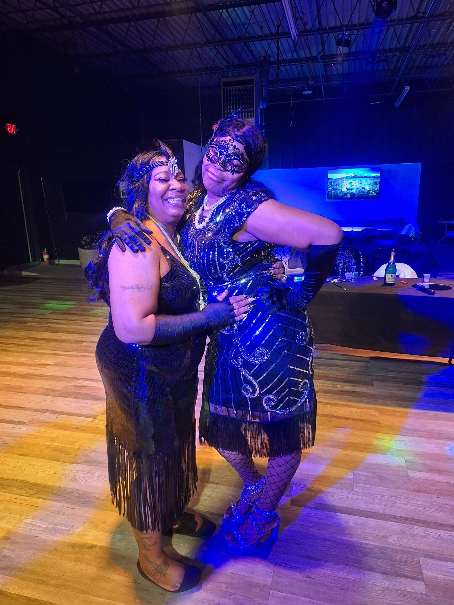 Two women in 1920s-inspired outfits hug and smile on a dimly lit dance floor, one wearing a lace mask and pearl necklace with fringed dress under blue stage lighting.