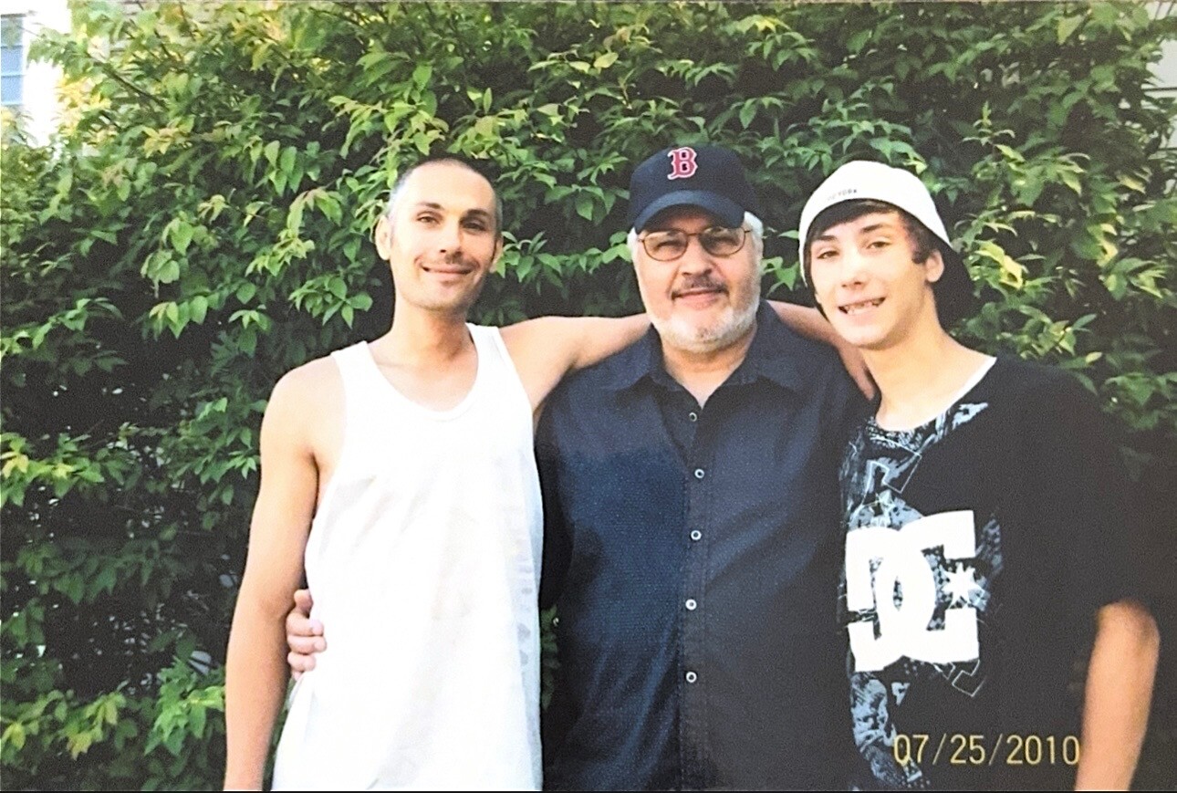 Three men pose outdoors with a leafy hedge behind them; left in a white tank top, middle in a dark shirt and glasses, right in a graphic tee and white cap. 07/25/2010 (date stamp)