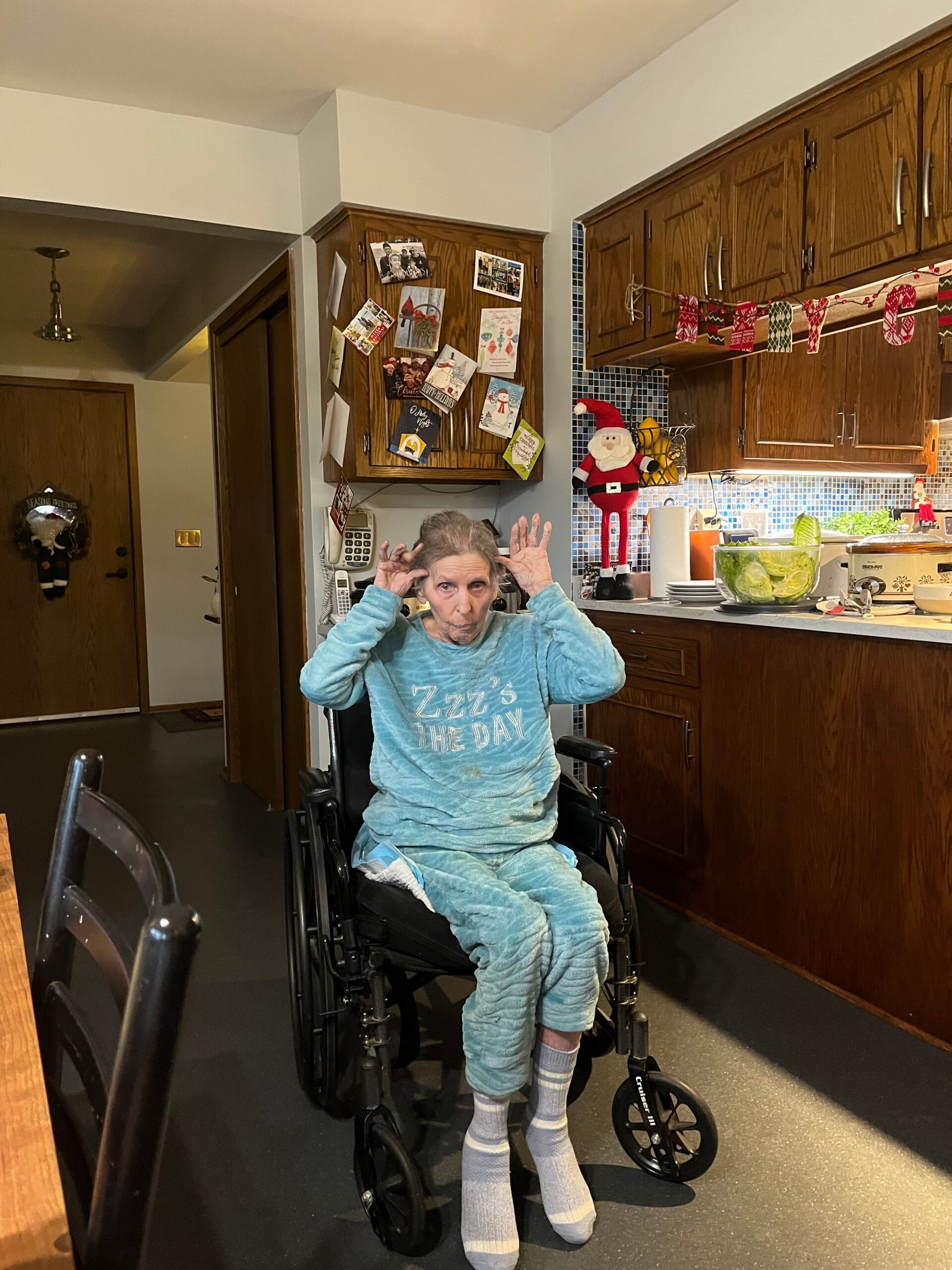 An elderly person in a wheelchair wearing a blue loungewear set, seated in a kitchen with holiday decorations on the cabinets and a Santa Claus figure nearby.