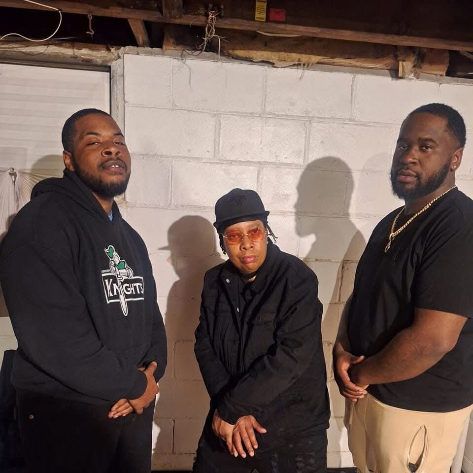Three adults posing indoors against a white brick wall; left man in a black Knights hoodie, center person in a black jacket and hat with orange-tinted sunglasses, right man in a black T-shirt and beige pants.