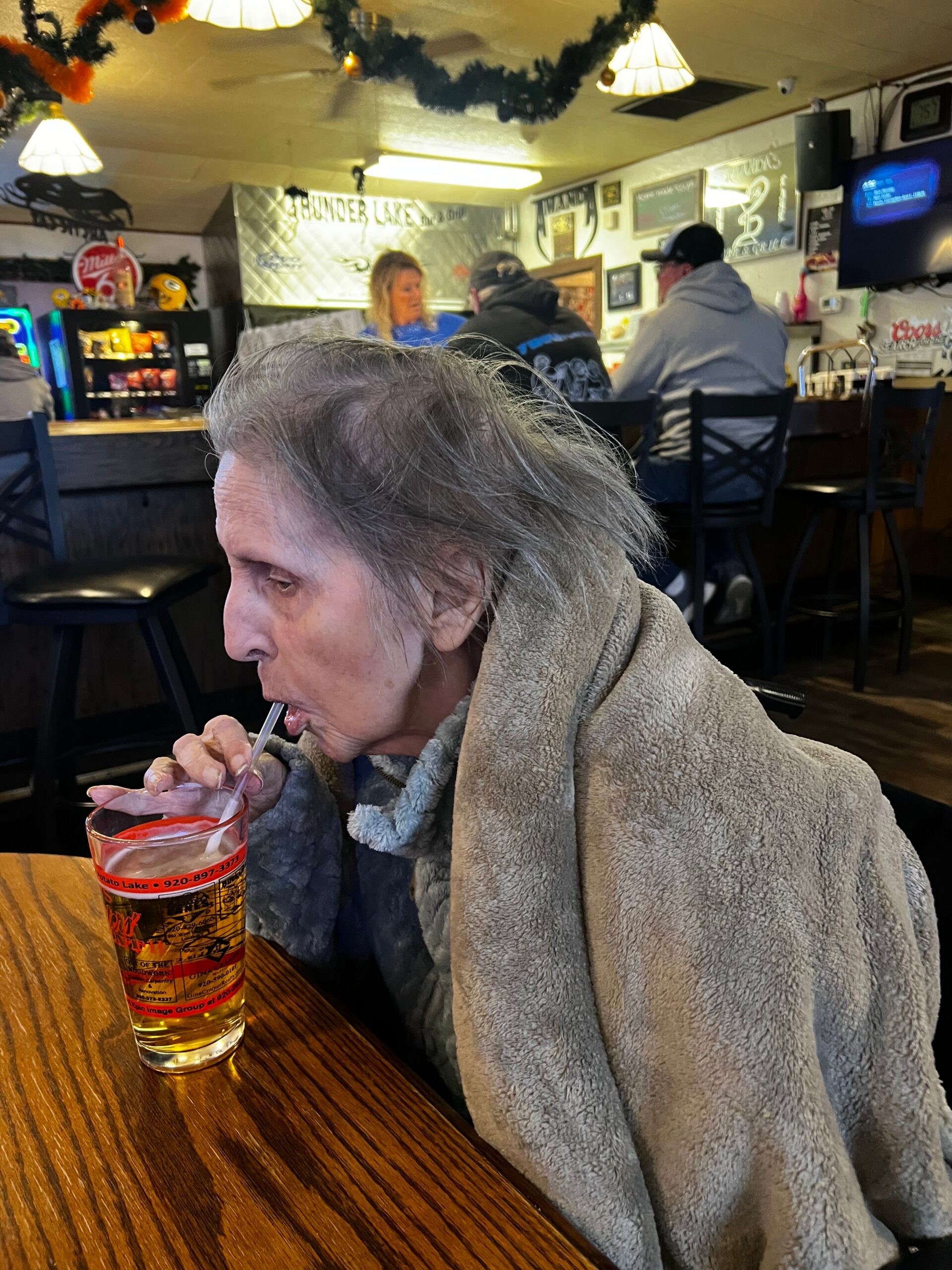 Elderly person with gray hair and a tan fleece blanket sits at a wooden bar, sipping a beer through a straw as people chat in the background.