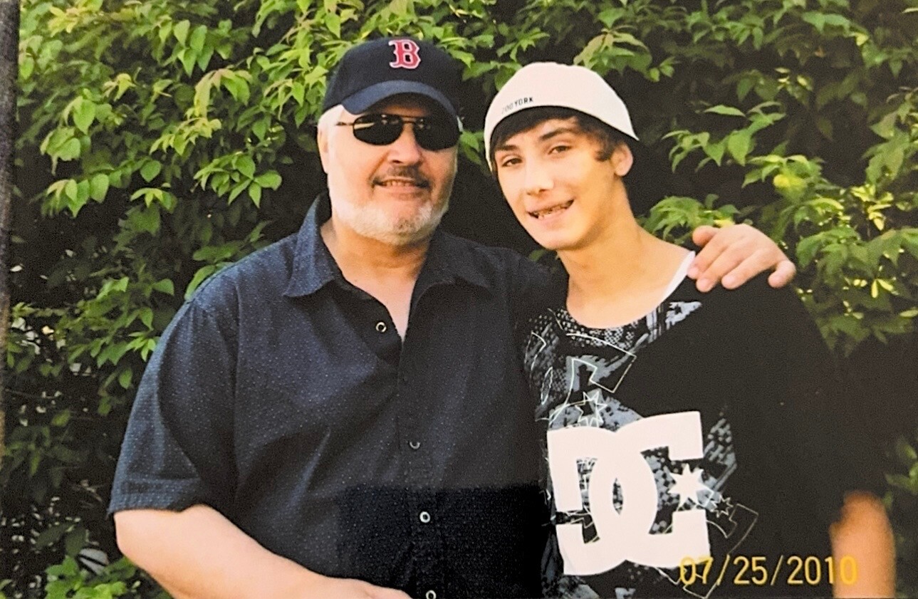Older man in a black cap with a red B and sunglasses stands with a younger man in a white cap, both smiling in front of green bushes.