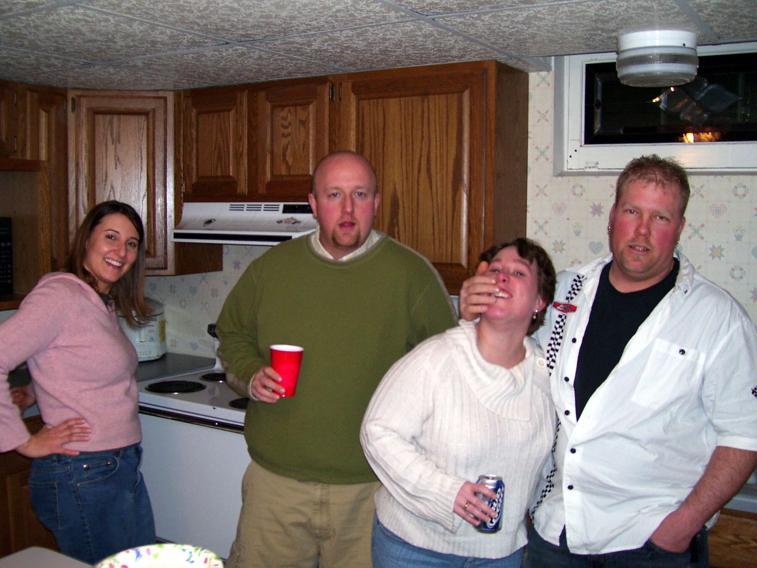 Group of four adults in a kitchen posing for the camera, one holding a red cup and another a drink can.