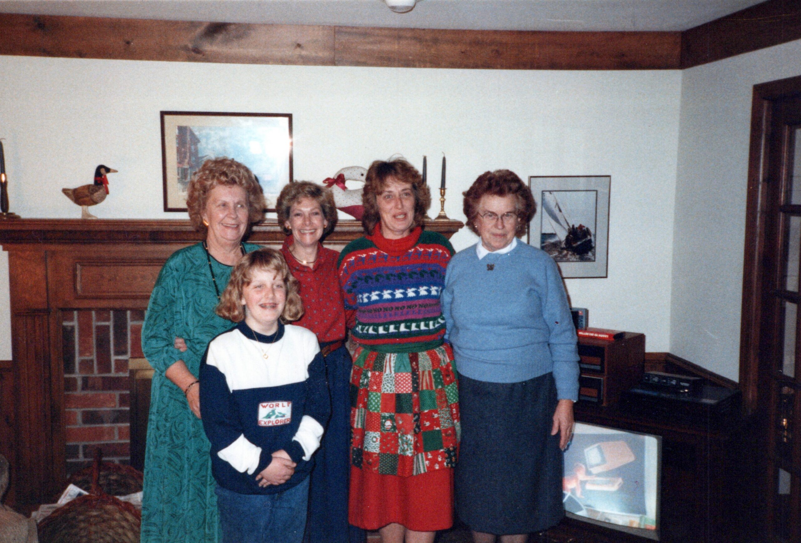 Group of five women and one girl posing together in a living room, wearing festive sweaters and smiling by a brick fireplace.