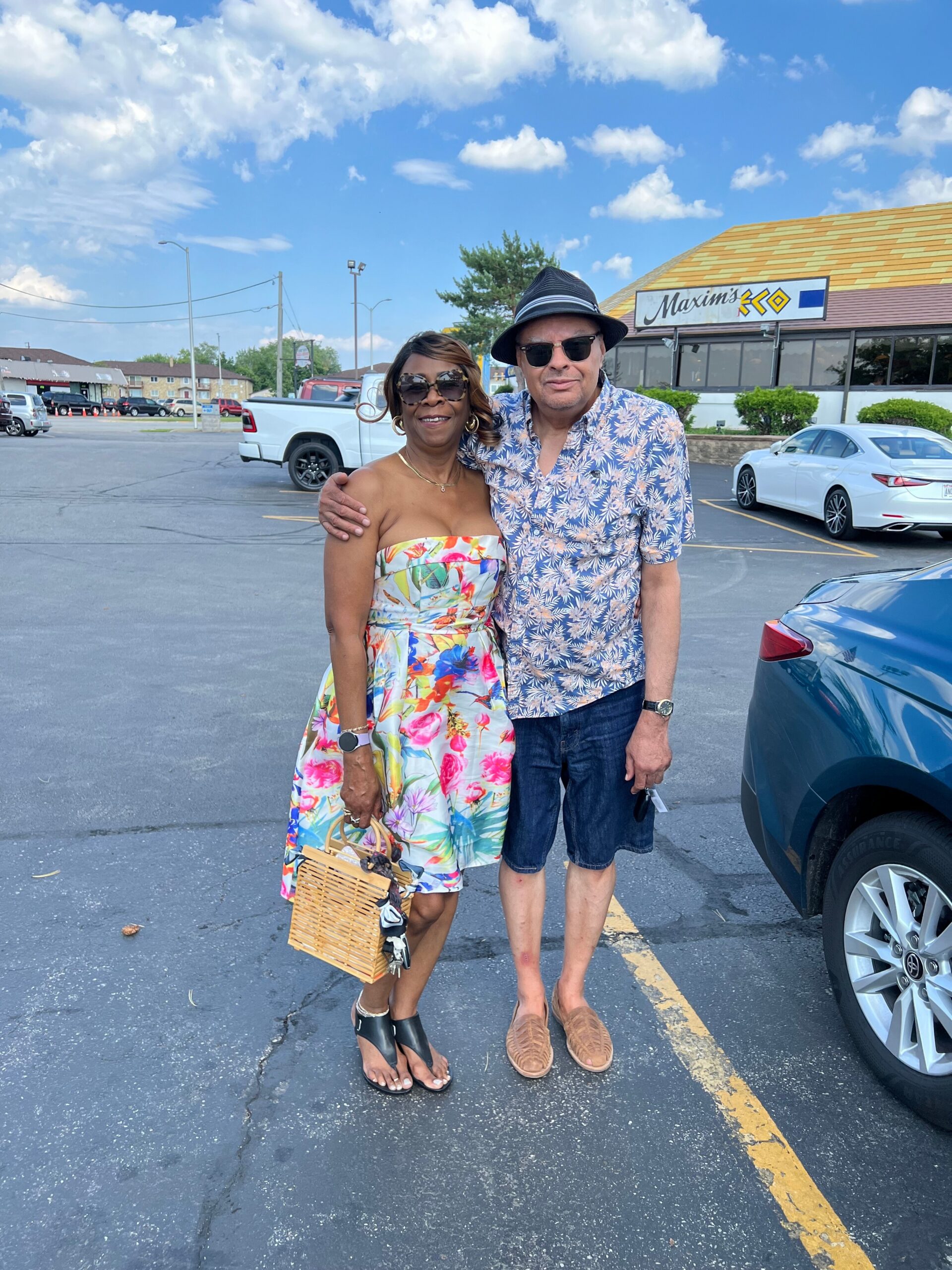 Couple posing together in a parking lot, woman in a colorful floral dress and man in a tropical shirt and hat, smiling at the camera with a basket resting by the woman