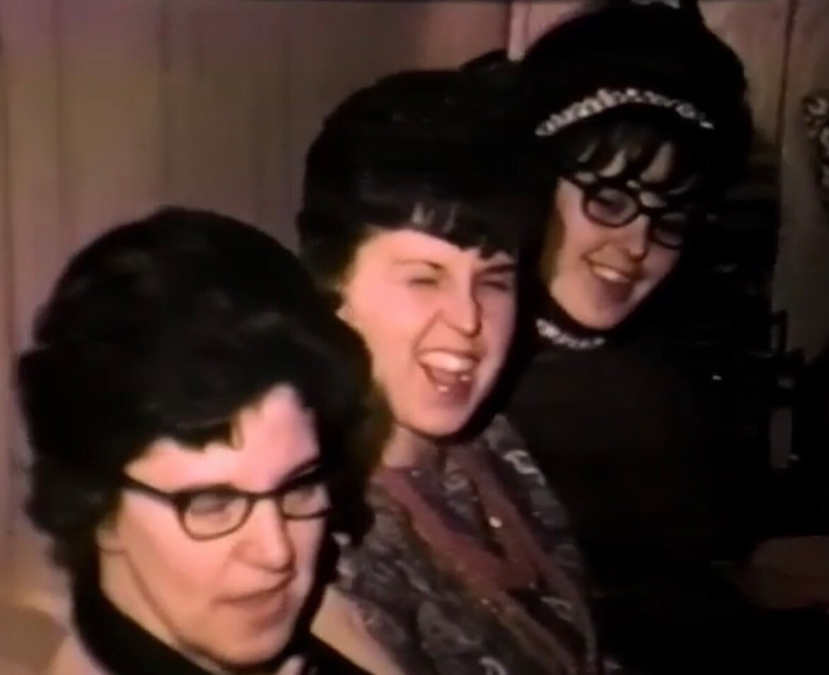 Three women in retro outfits laugh together at a casual gathering, smiling broadly at the camera inside a dim room.